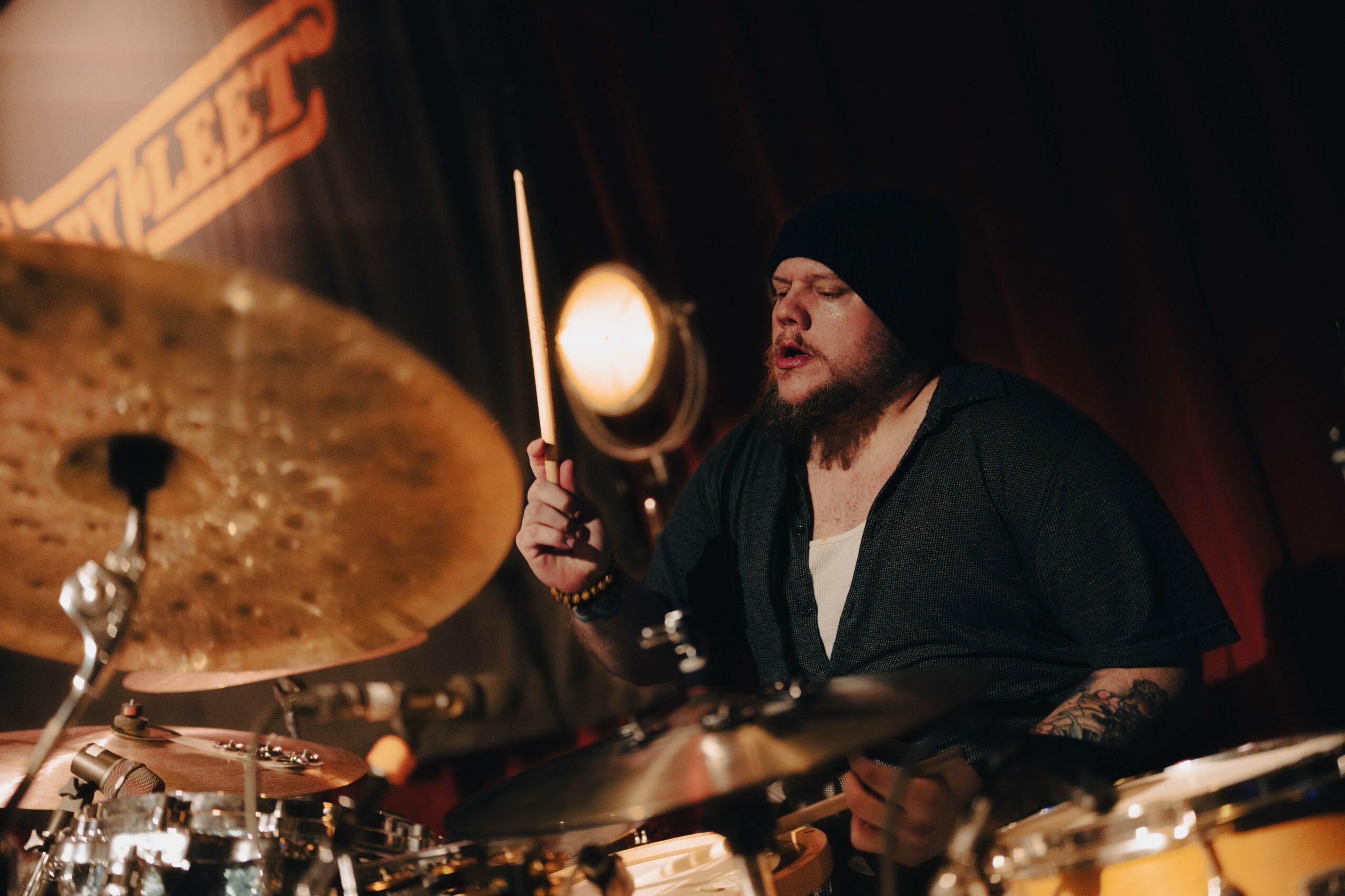 Bearded drummer wearing a black beanie and shirt, playing drums on stage with a cymbal nearby.