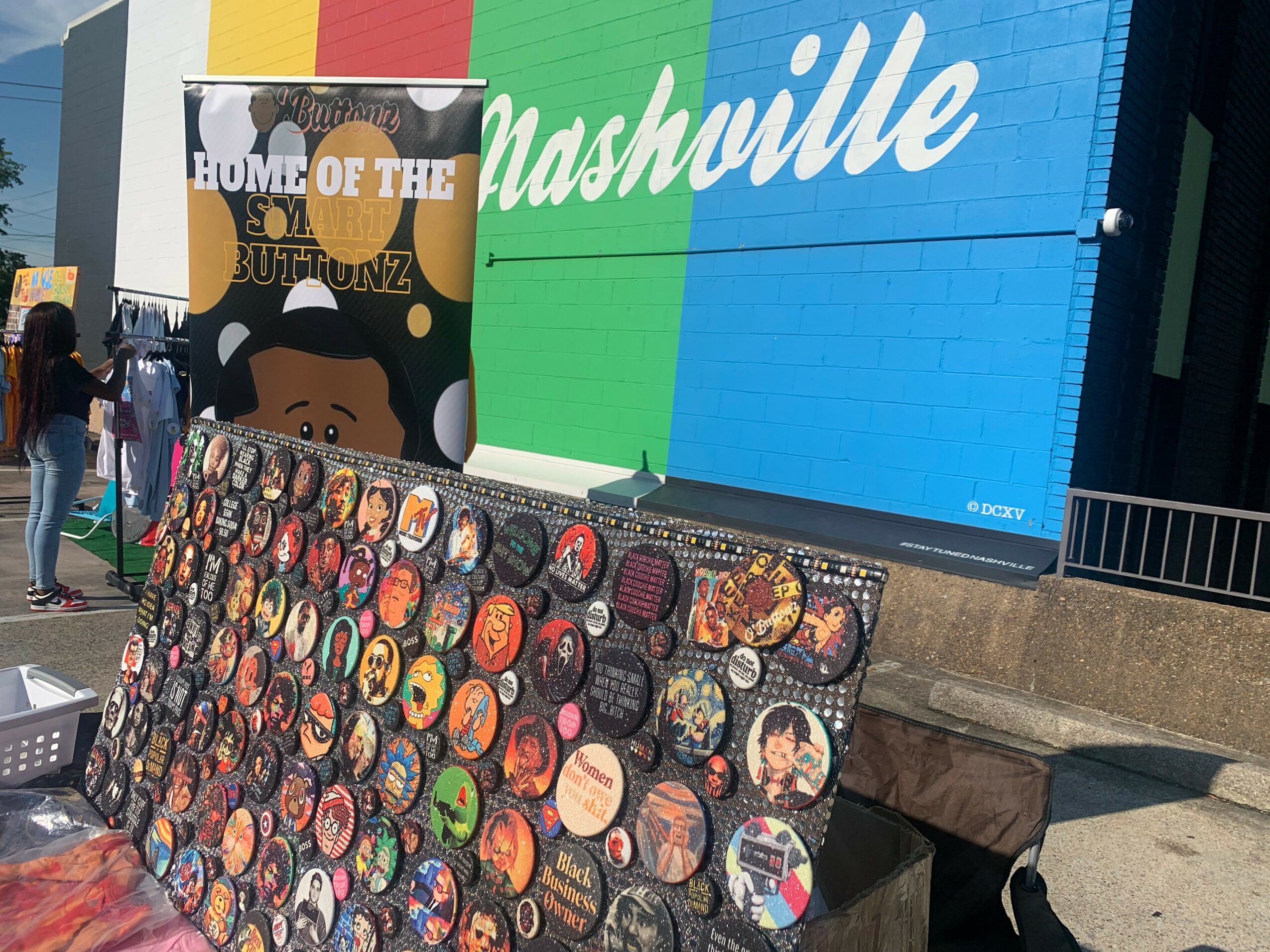 Outdoor market stall with colorful badges and a Nashville sign in the background.