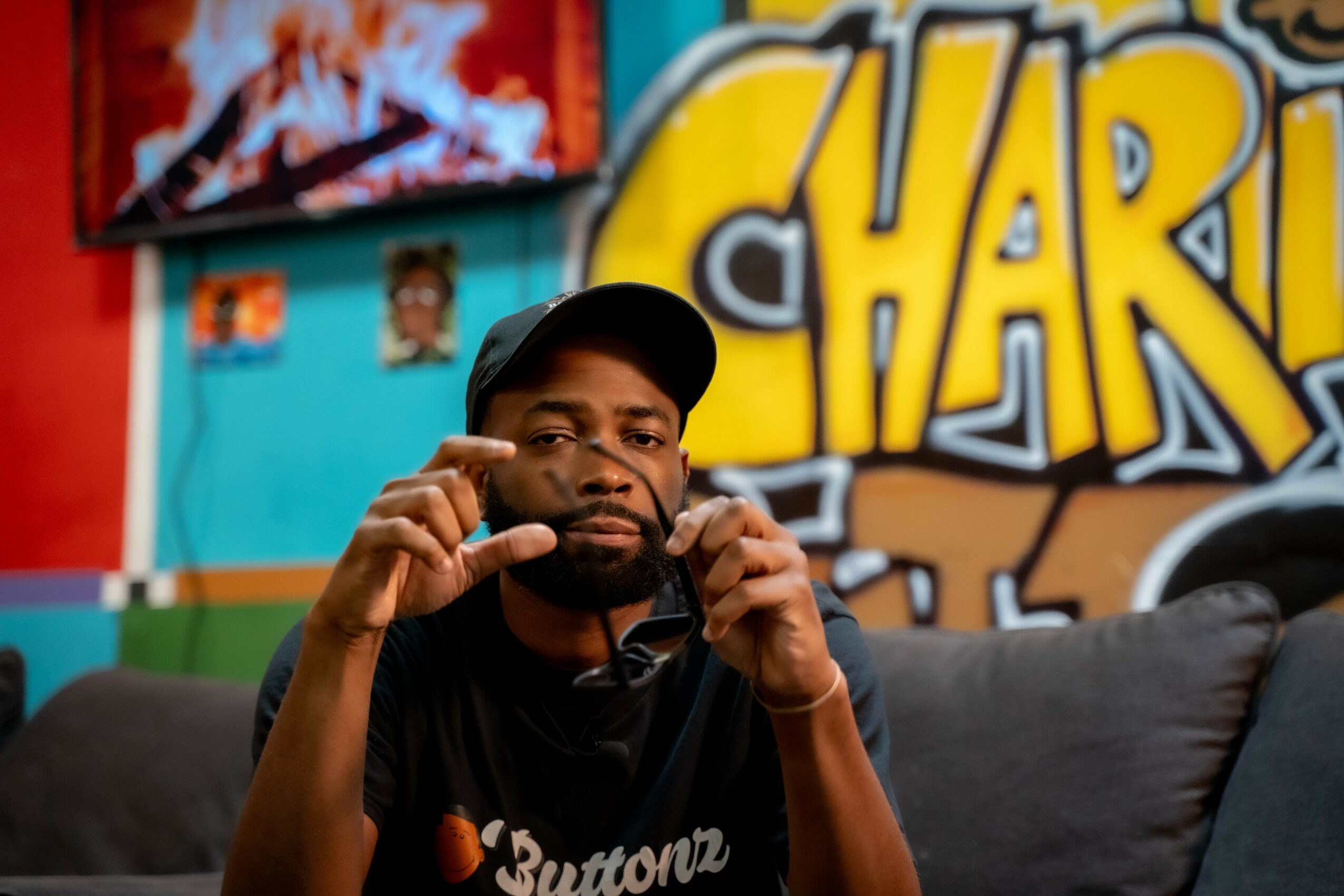Man with beard and cap sitting on a couch in a colorful room with graffiti art on the wall behind him.