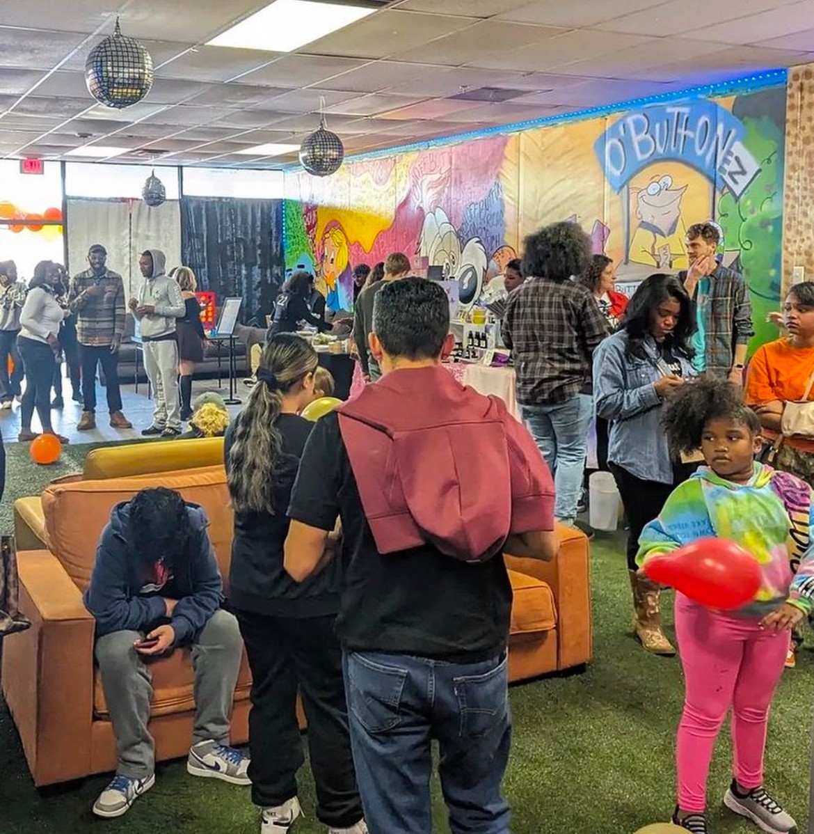 People gathered in an indoor space with colorful wall art and decorations, some sitting on couches, others standing and talking.