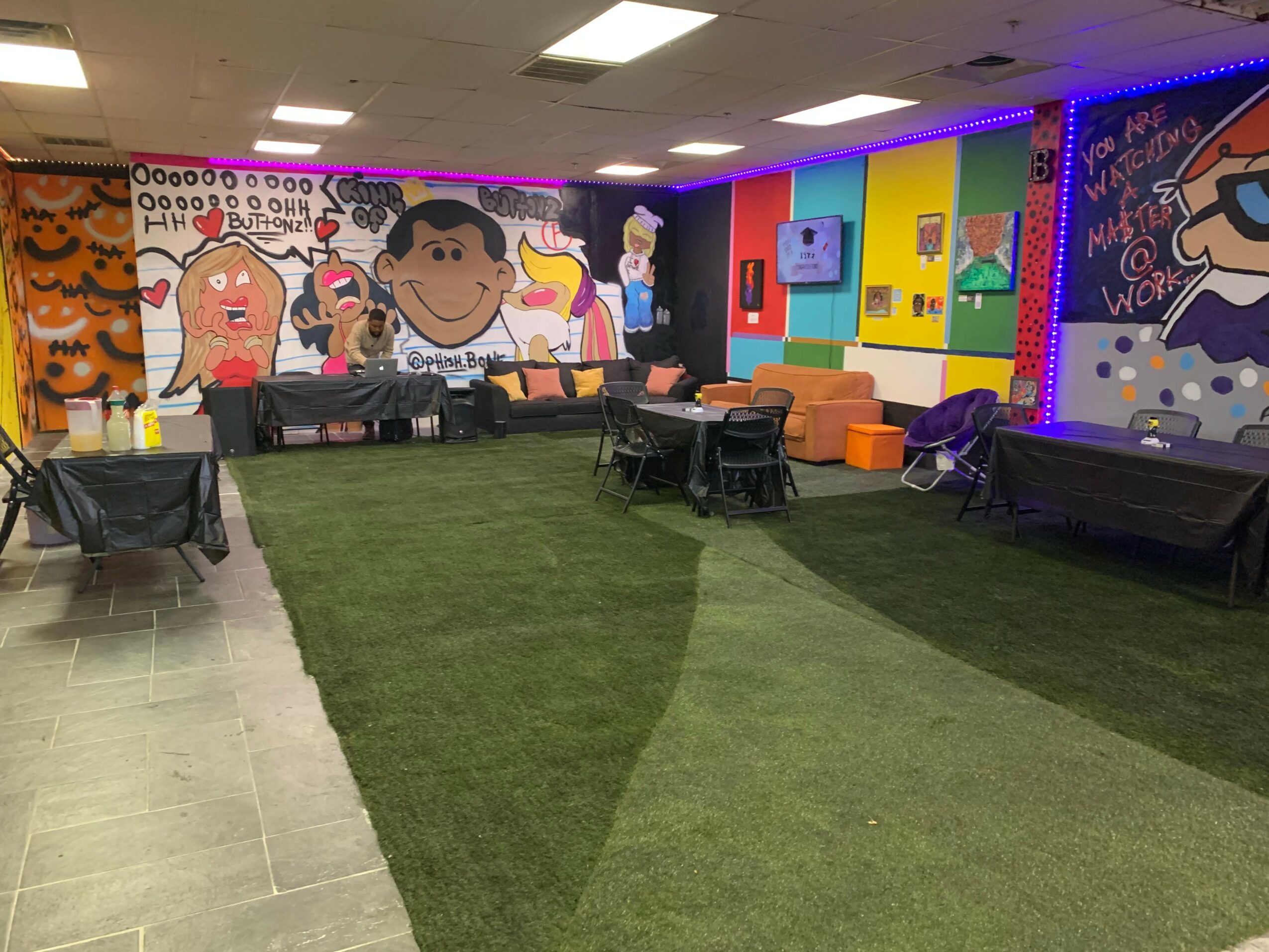 Indoor space with colorful wall murals, black tables, orange seating, and artificial grass flooring, illuminated by ceiling lights.