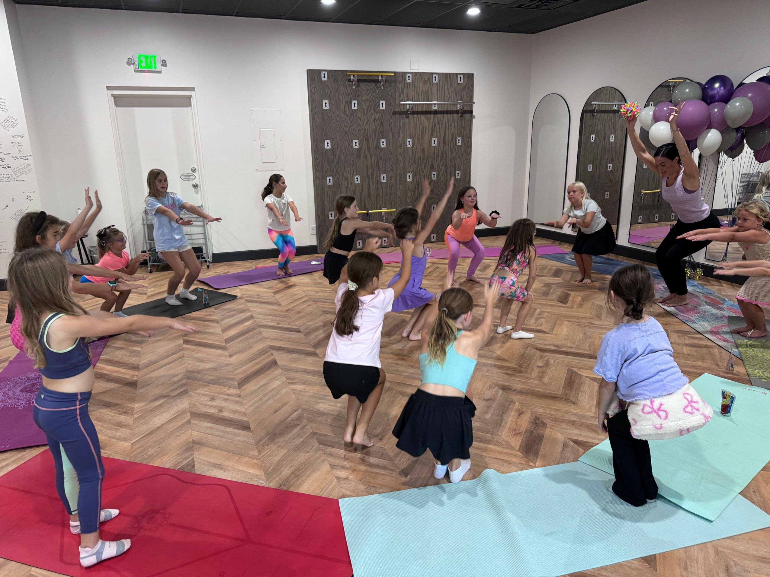 Group of children participating in a fitness class led by an instructor in a room with balloons and wooden flooring.