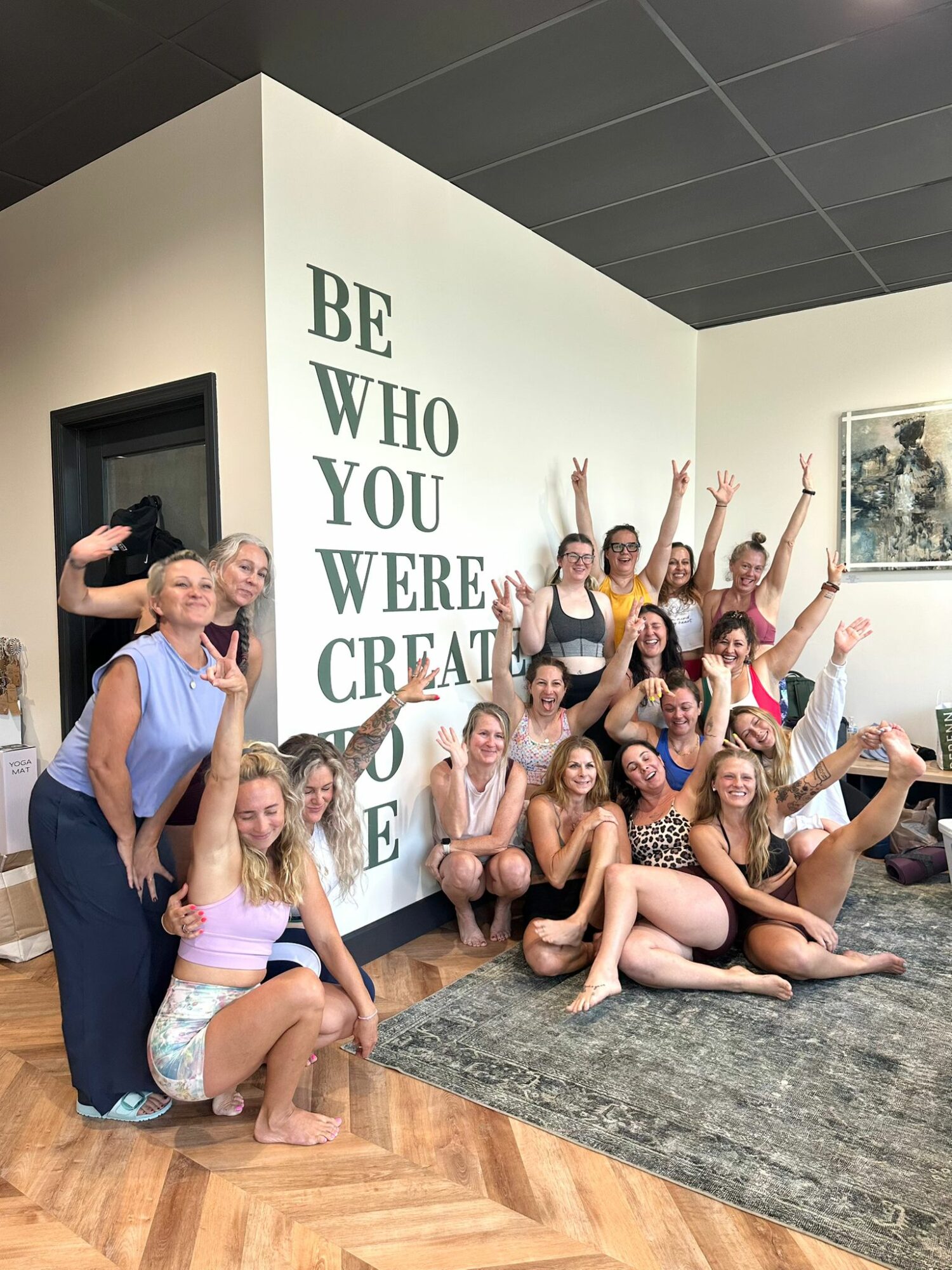 Group of young women posing indoors, some sitting and others standing with arms raised, in front of a wall with motivational text.