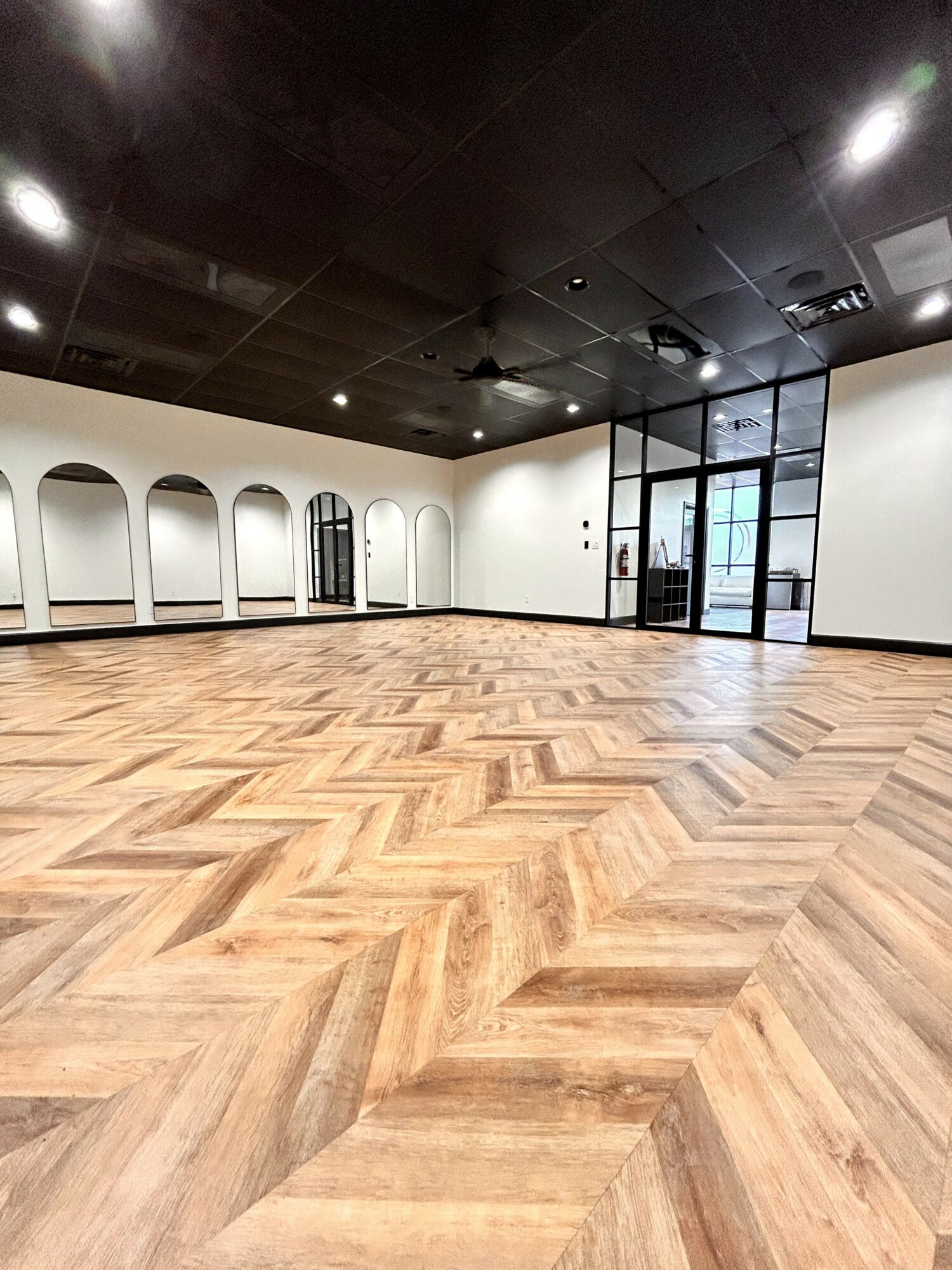 Empty room with wooden herringbone floor, white walls, black ceiling, and large glass doors and windows.
