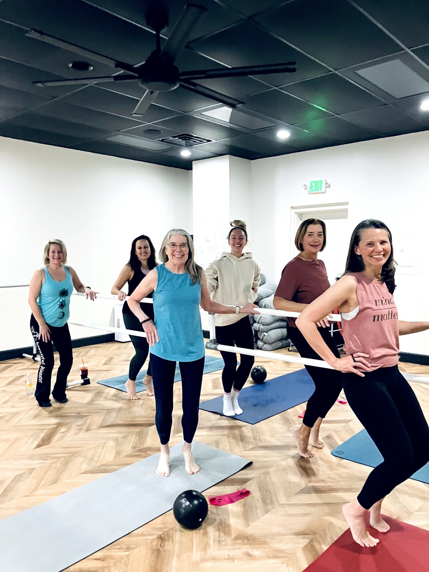 Group of women in a fitness class standing on mats in a room with white walls and wooden floor.