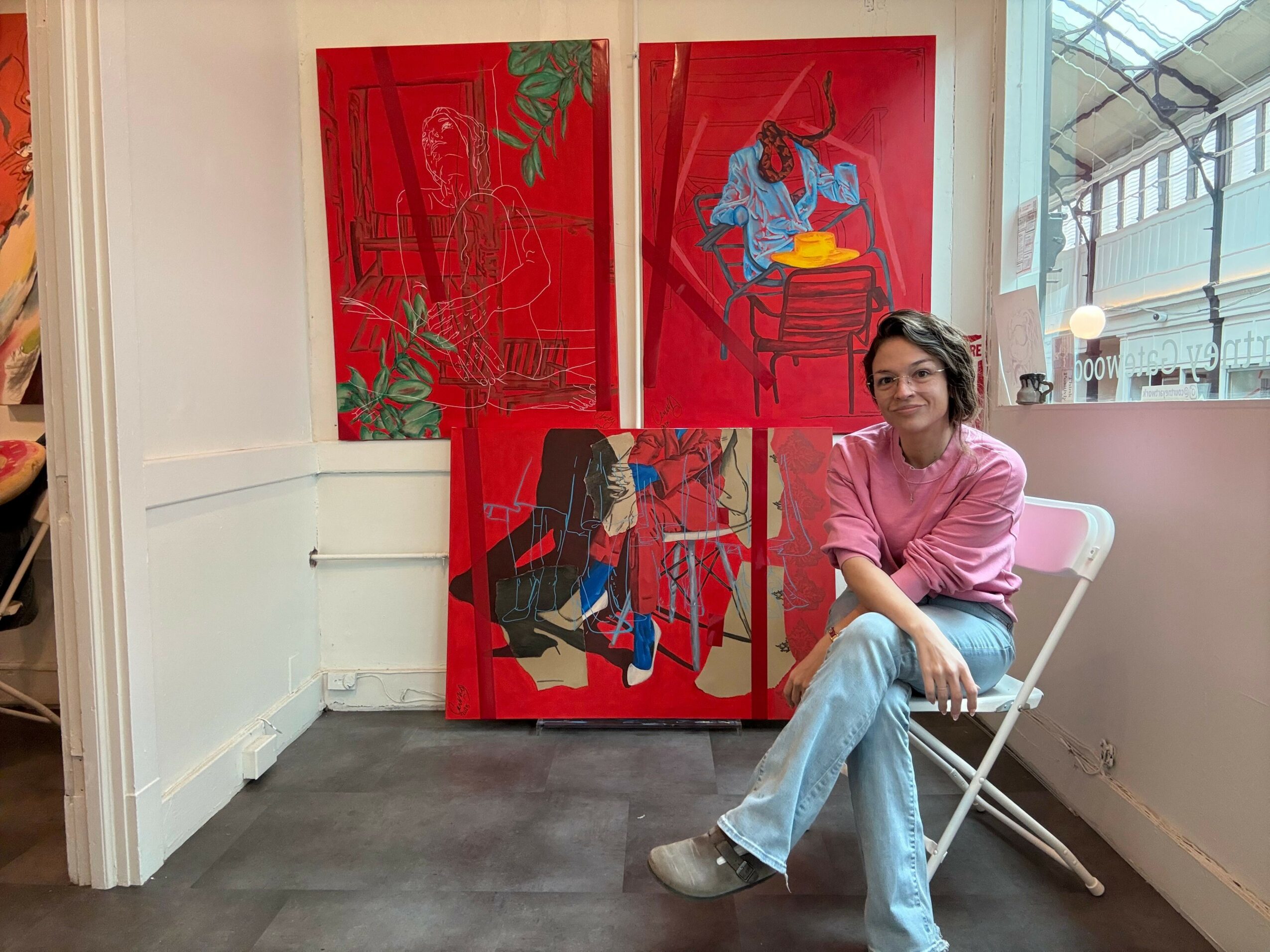 Woman sitting on a white folding chair in front of three colorful paintings on a wall, with a window to the right.