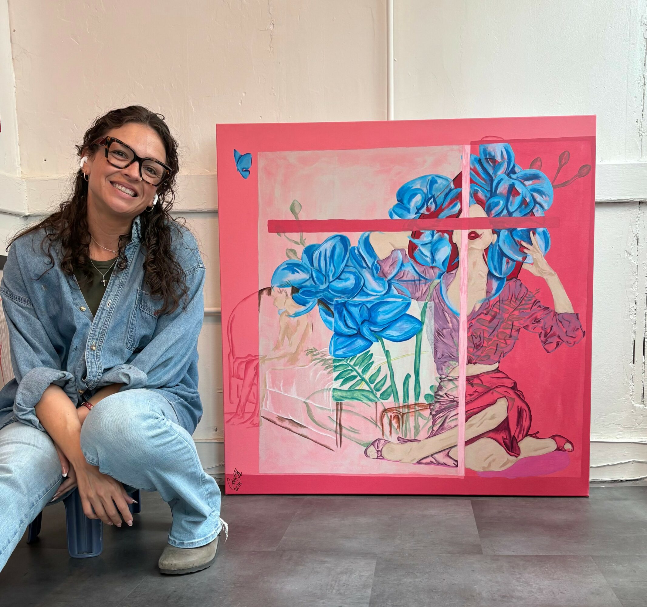Woman with glasses and curly hair sitting next to a colorful painting of blue flowers and a person in pink background.