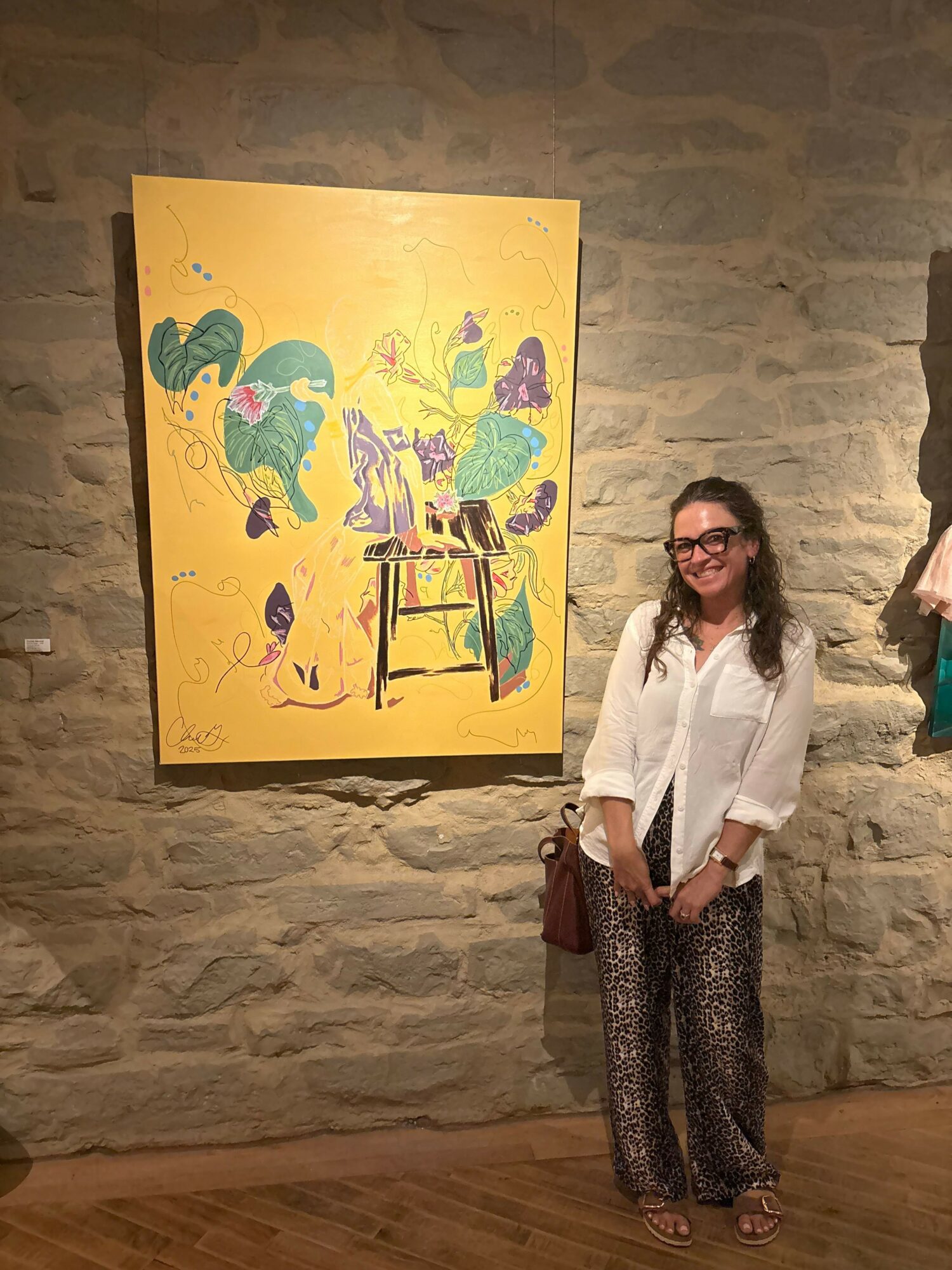 Woman with glasses and long hair standing next to colorful artwork on stone wall, smiling, wearing white top and patterned pants.