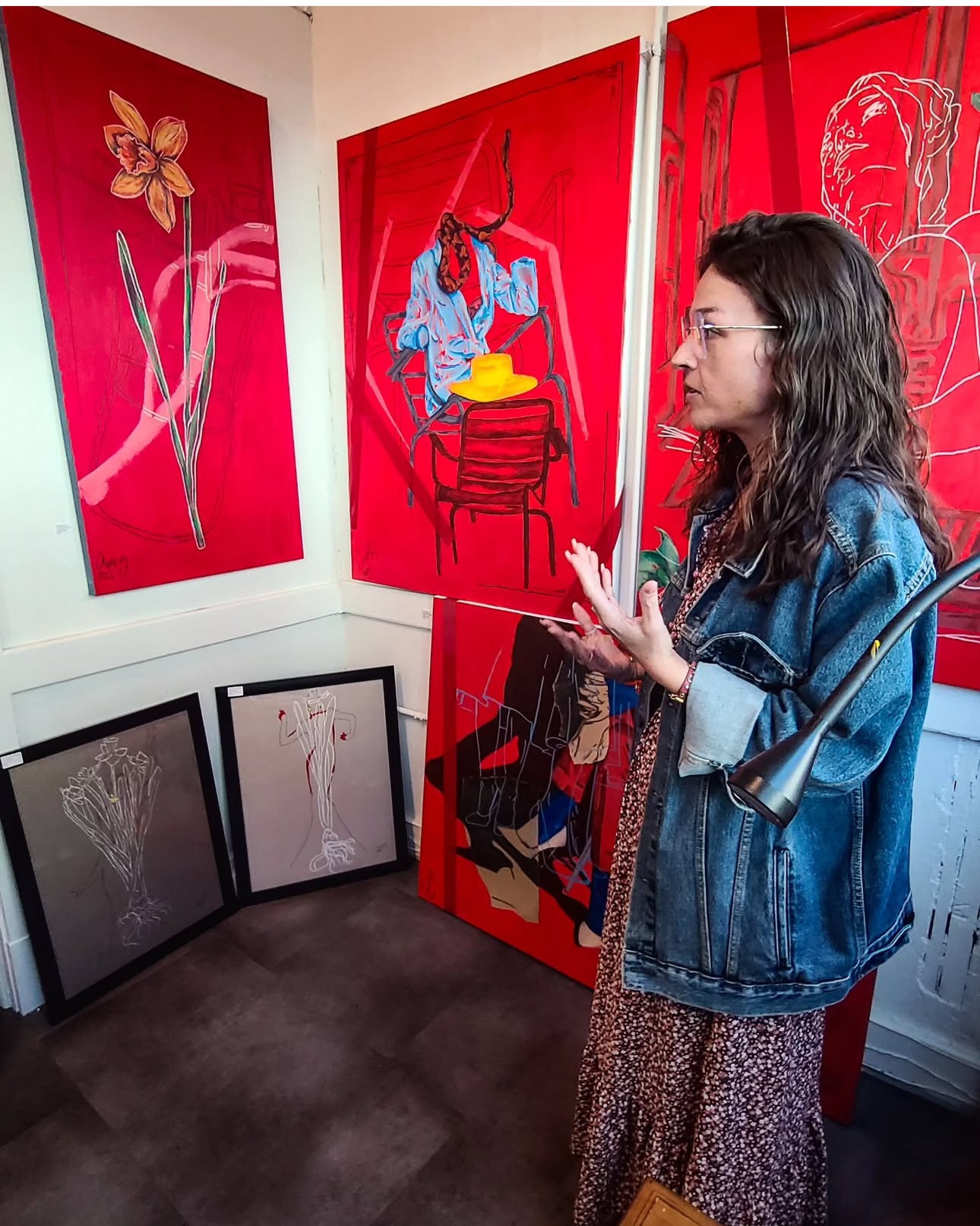 Woman with glasses and curly hair wearing a denim jacket and patterned dress looking at red paintings in an art gallery.