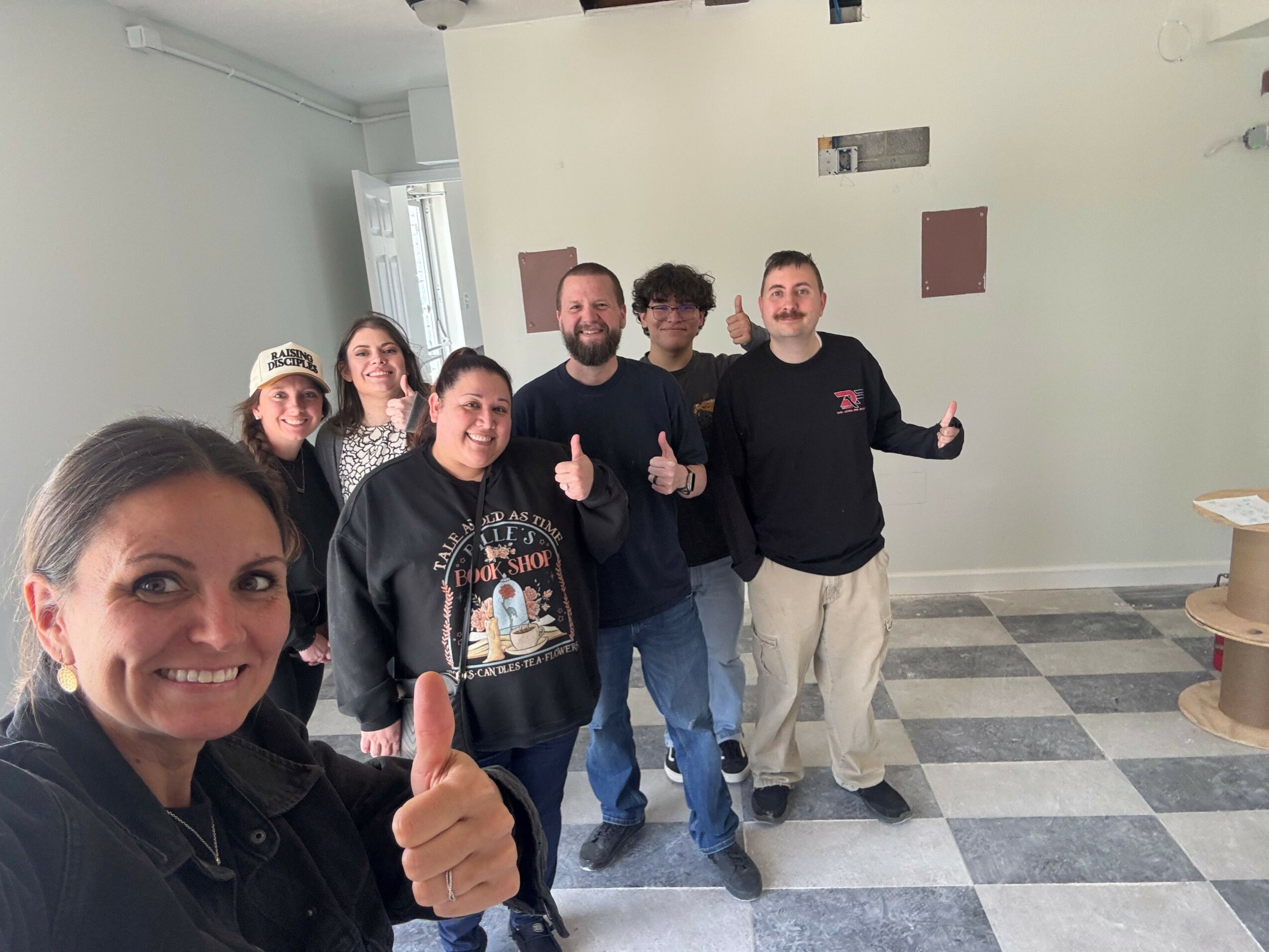 Group of six people standing indoors, smiling, some giving thumbs-up, with a checkered floor and plain wall background.