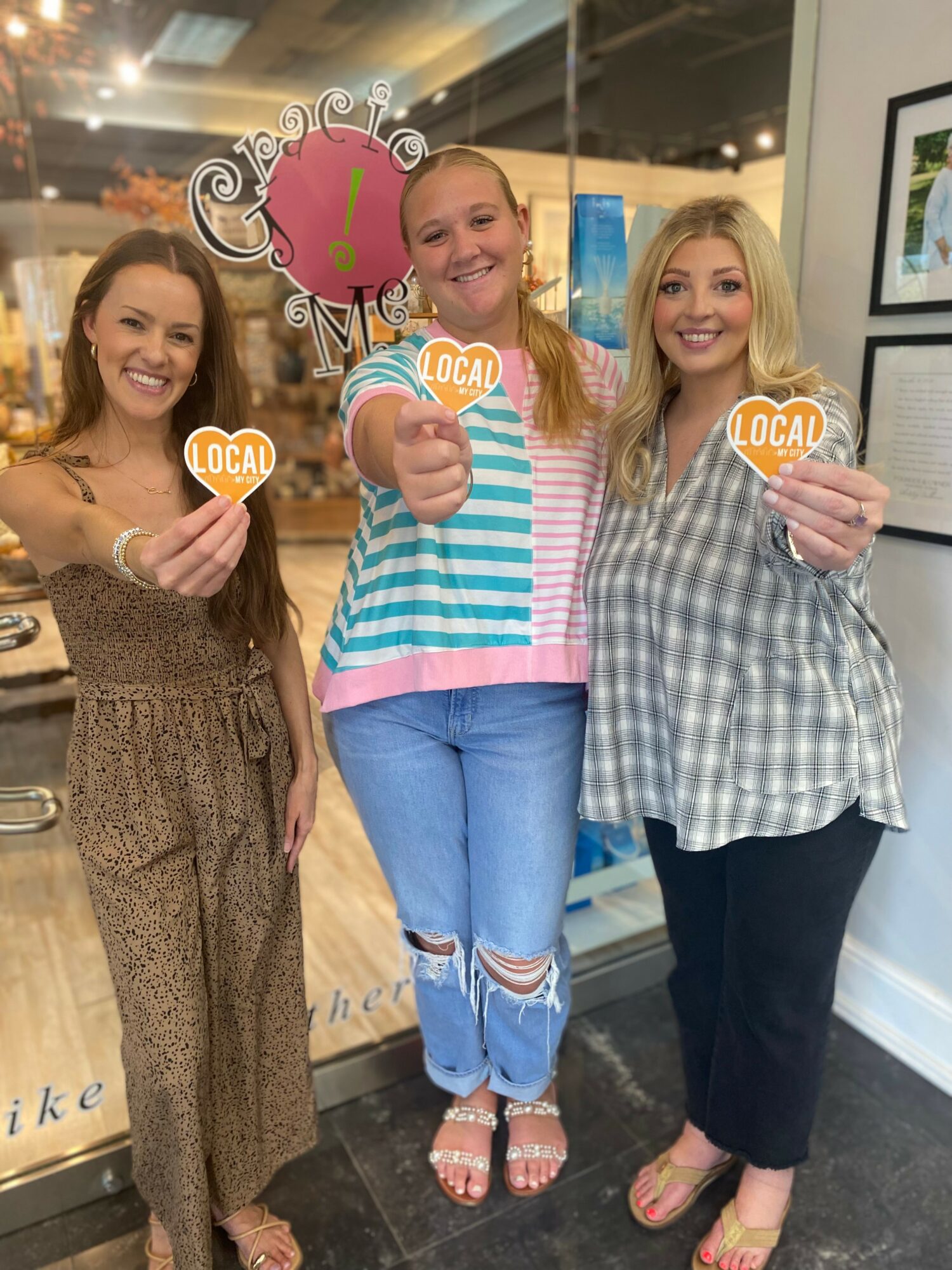 Three women holding heart-shaped cookies with 'local' written on them, smiling indoors.