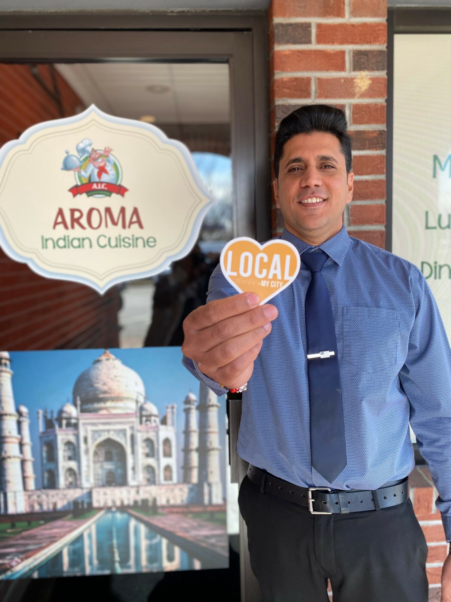 Man in blue shirt and tie holding a 'Local' sign outside a restaurant with an Indian cuisine sign and Taj Mahal picture.