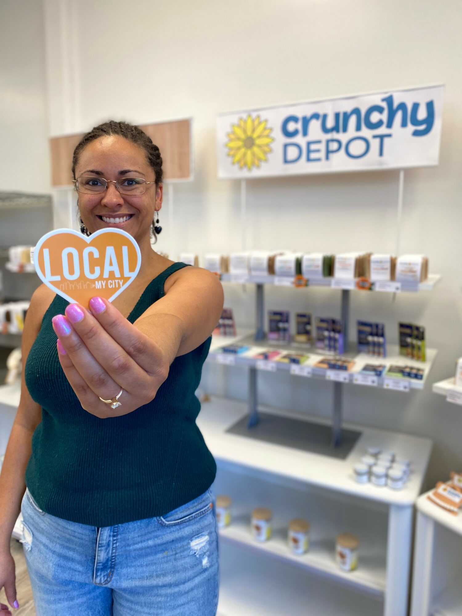 Woman smiling and holding a heart-shaped sign that says 'LOCAL' in front of a store display.