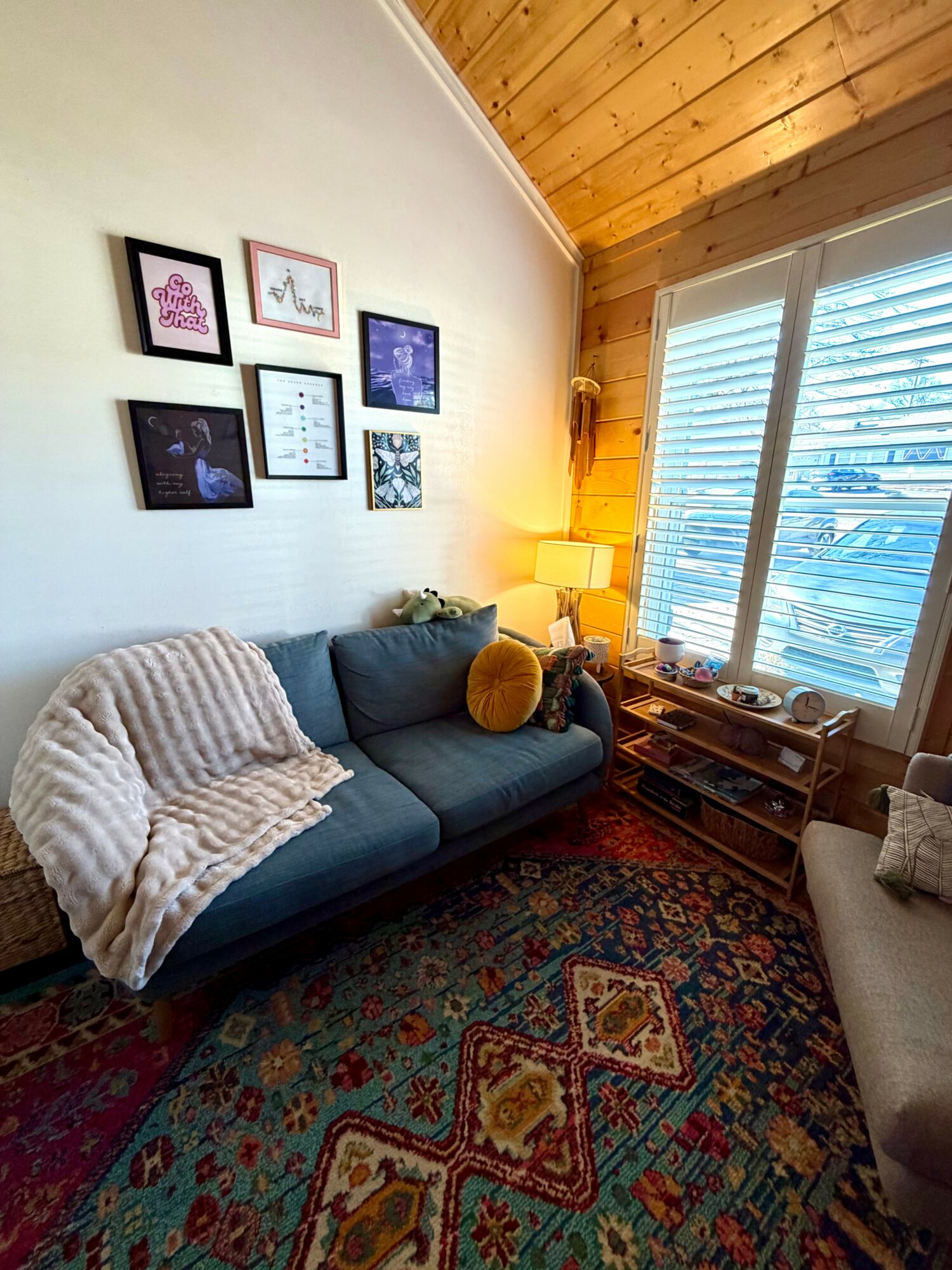 Living room with a dark sofa, beige blanket, wall art, wooden ceiling, window with blinds, and a patterned rug.