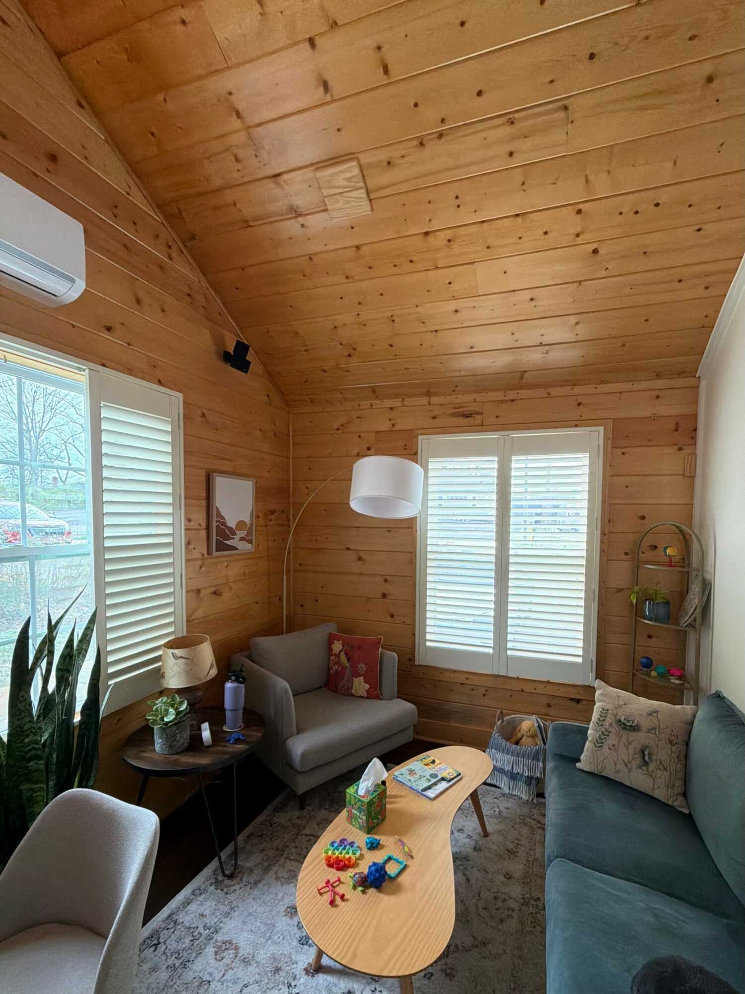 Living room with wooden walls and ceiling, sofa, armchair, coffee table, and windows with blinds.