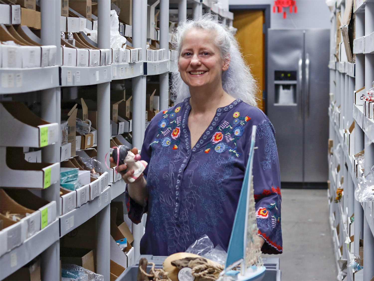 Smiling woman with gray hair in a blue embroidered blouse holding a small object in a storage room with shelves of boxes.
