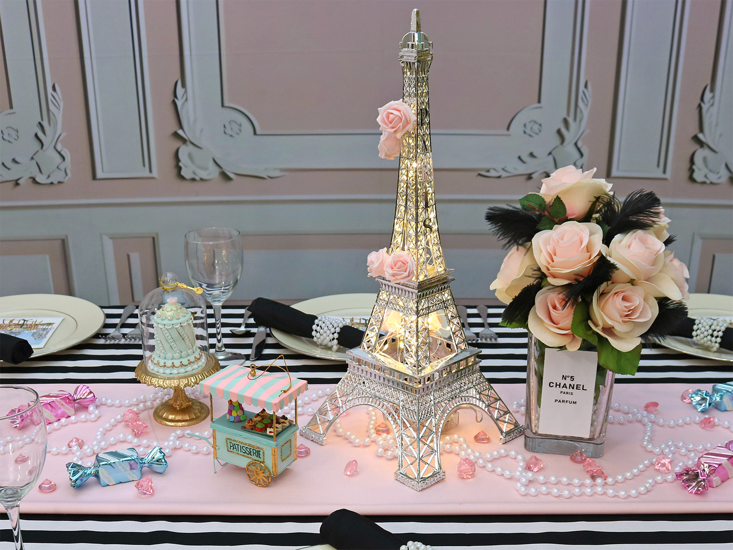 Decorative table with Eiffel Tower centerpiece, pink roses, jewelry, and small gift boxes, set against a pastel wall background.