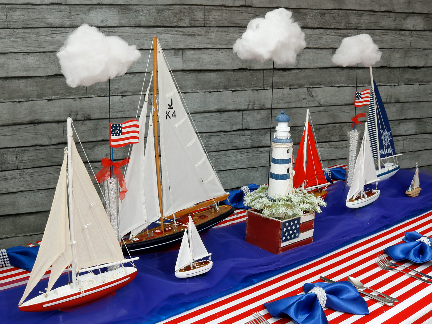 Model sailboats, lighthouse, clouds, and American flags on a striped tablecloth with a wooden background.