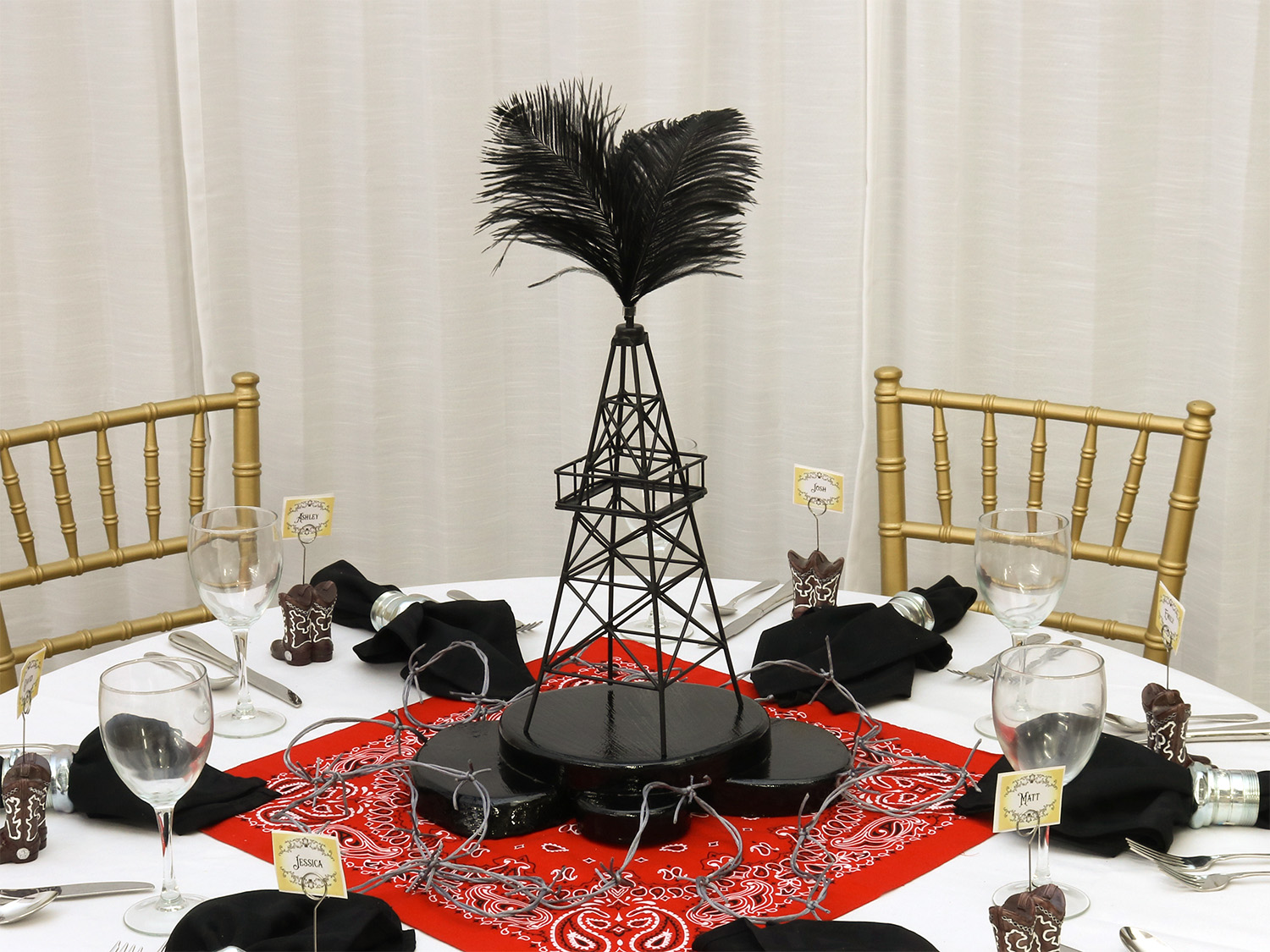 Table setting with gold chairs, a red patterned tablecloth, black napkins, glasses, and a centerpiece with black feathers.