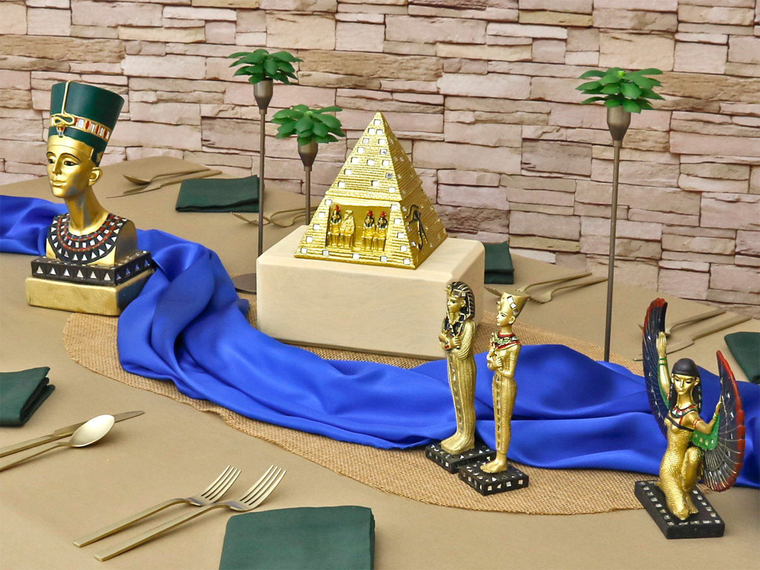 Decorative Egyptian-themed display with statues, a pyramid, and blue fabric on a table, against a brick wall background.