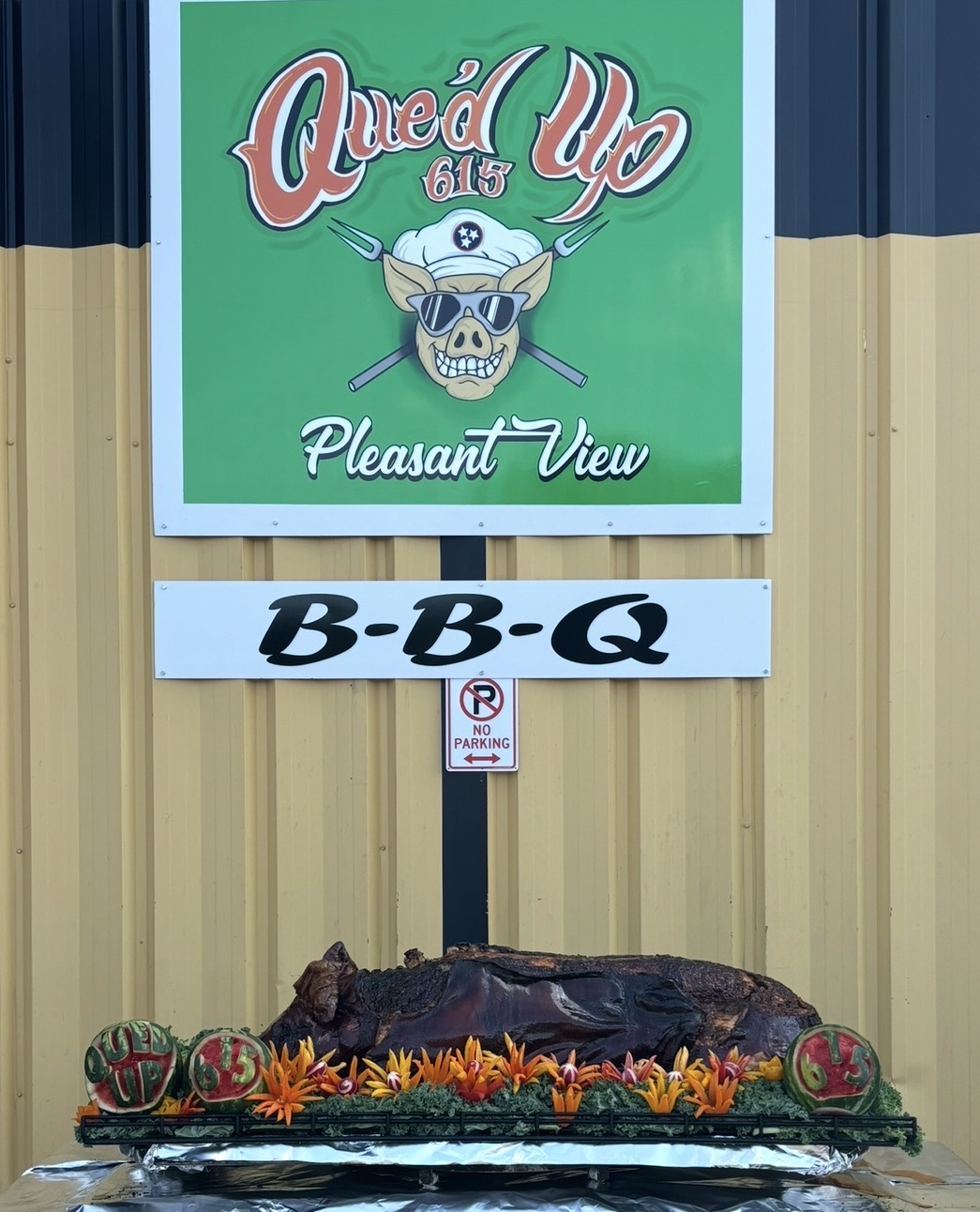 Sign with a skull wearing a sailor hat, text 'Que'd Up', and a barbecue display below with decorative pumpkins and flowers.