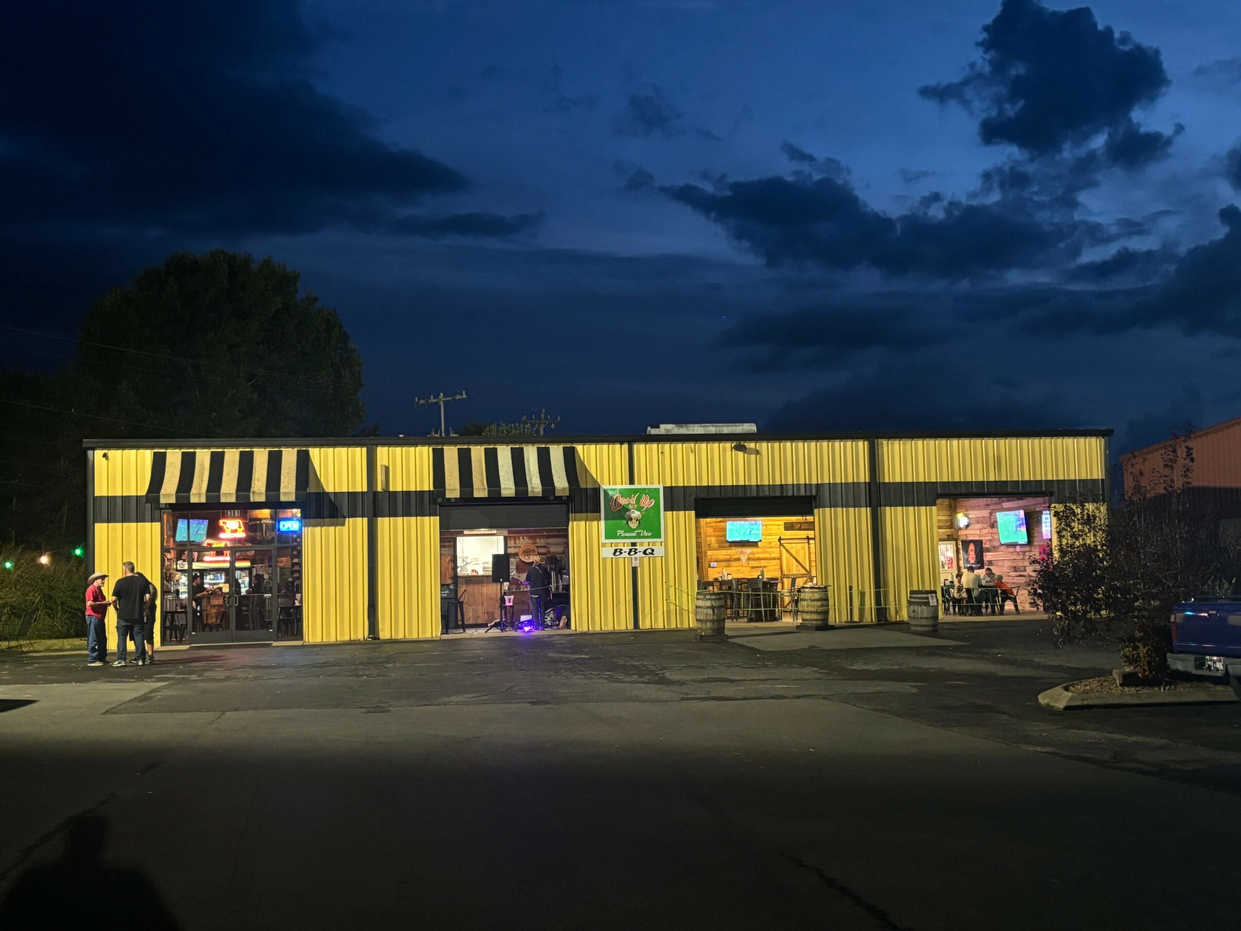 Yellow building with black and yellow striped awning, night sky with dark clouds, people standing outside, parking lot in front.