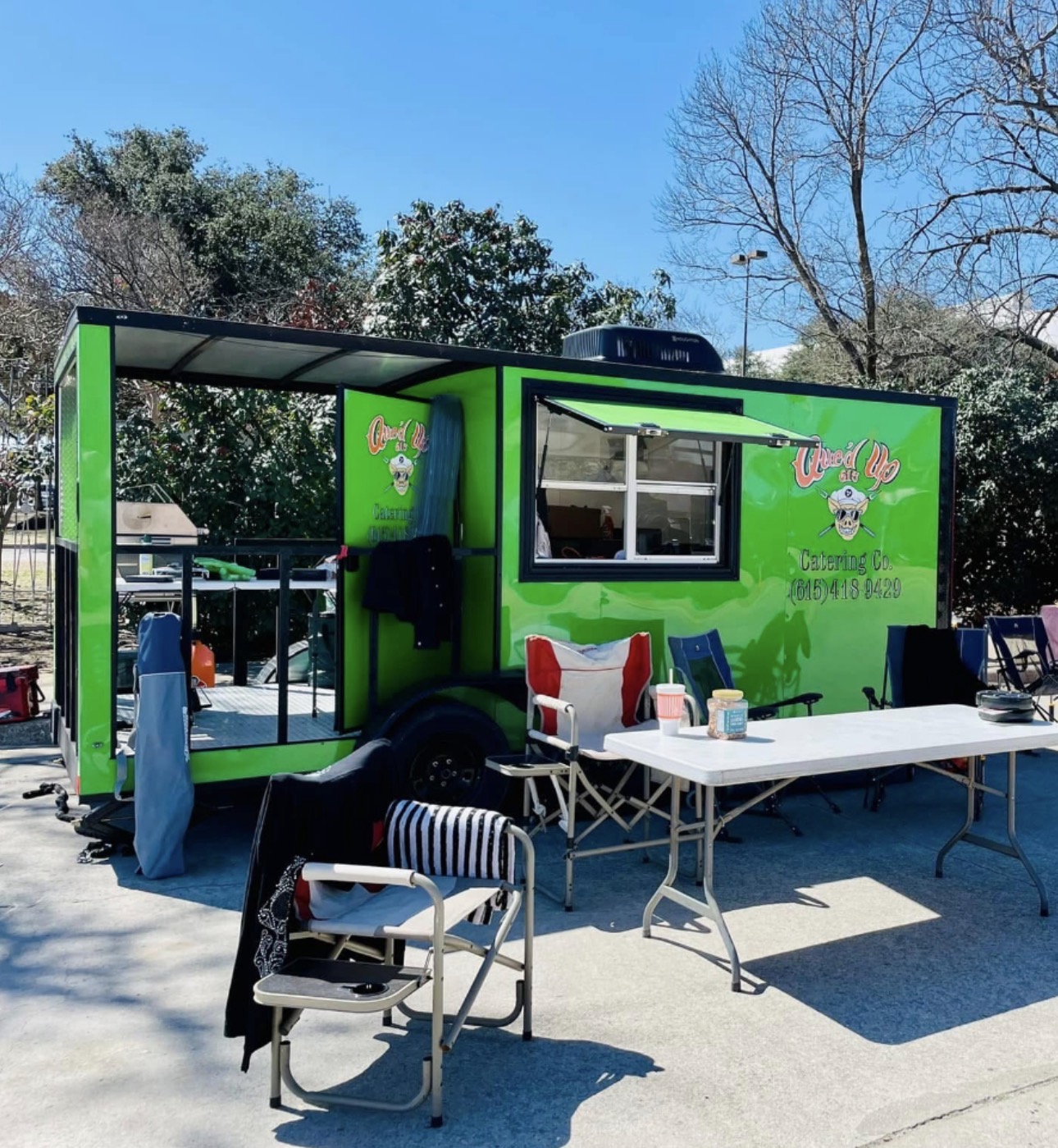 Green food truck with a window, outdoor table with chairs, and trees in background.