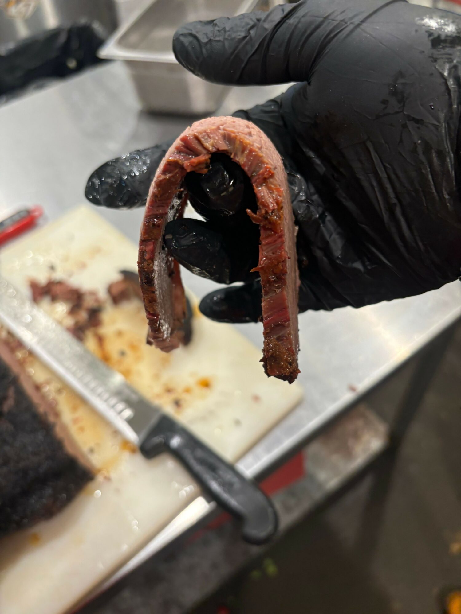Person wearing black glove holding a cooked, curved sausage over a kitchen counter with a cutting board and knife.