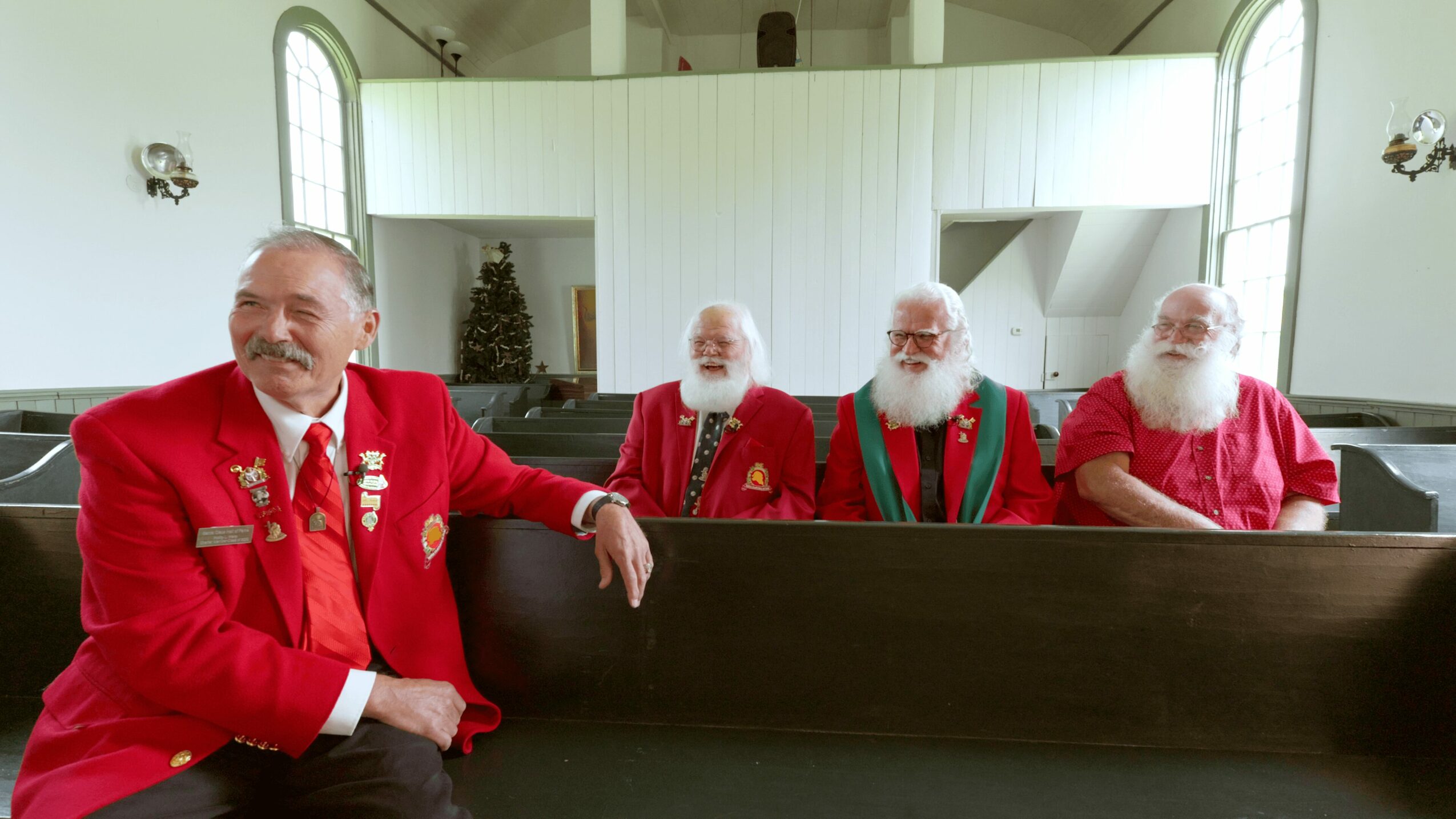 Four men with white beards and red clothing sitting in a church pew inside a bright room with arched windows.