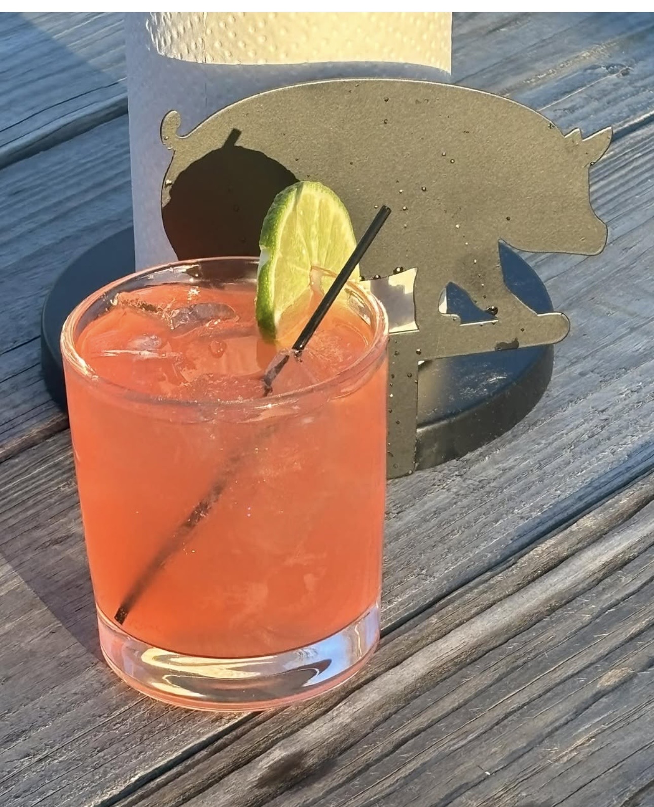 Pink cocktail with lime wedge and straw on wooden table, shadow of pig-shaped cutout behind it.