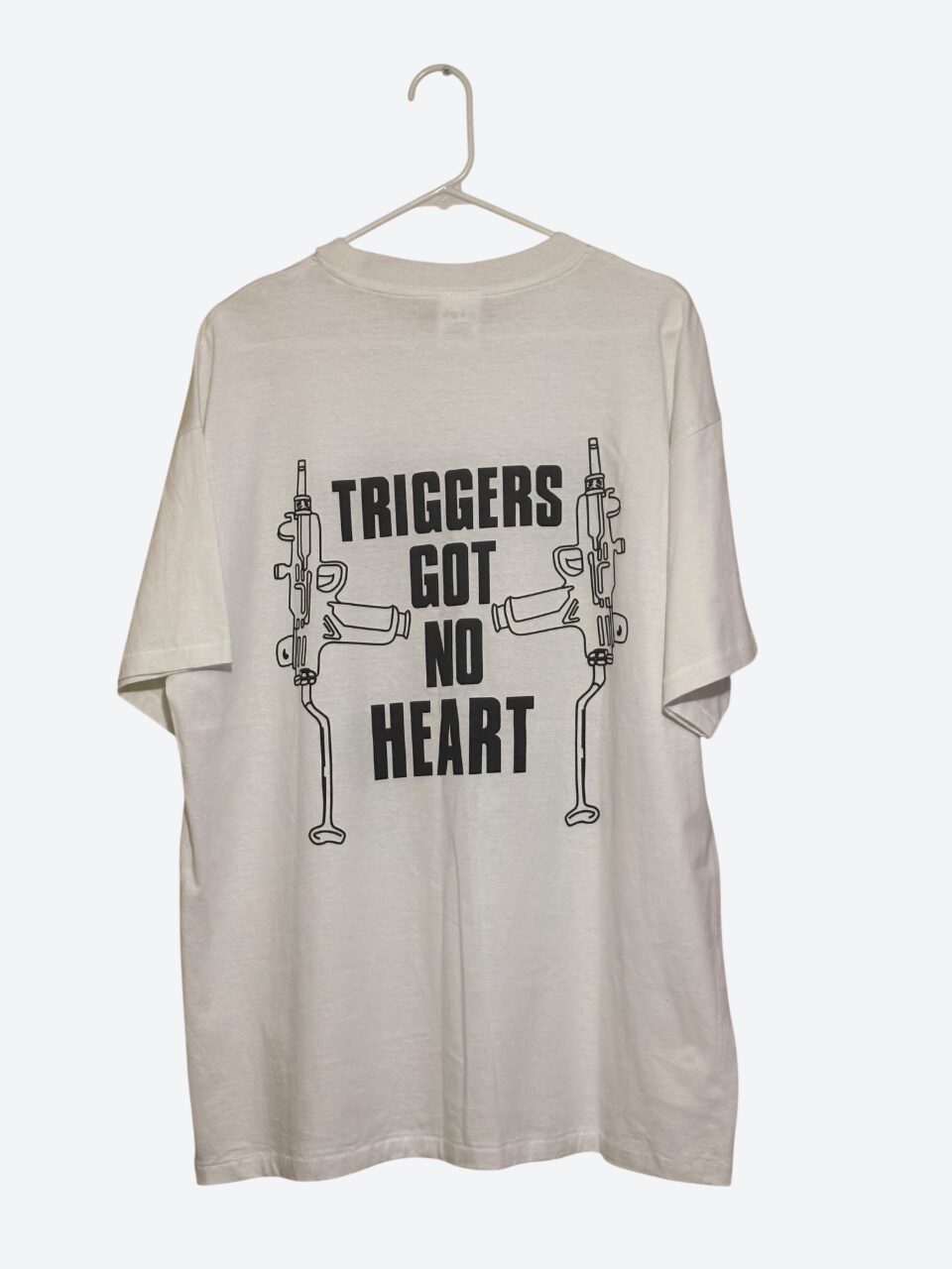 T-shirt with graphic of guns and text 'Triggers Got No Heart'. Hanging on a hanger against a plain background.