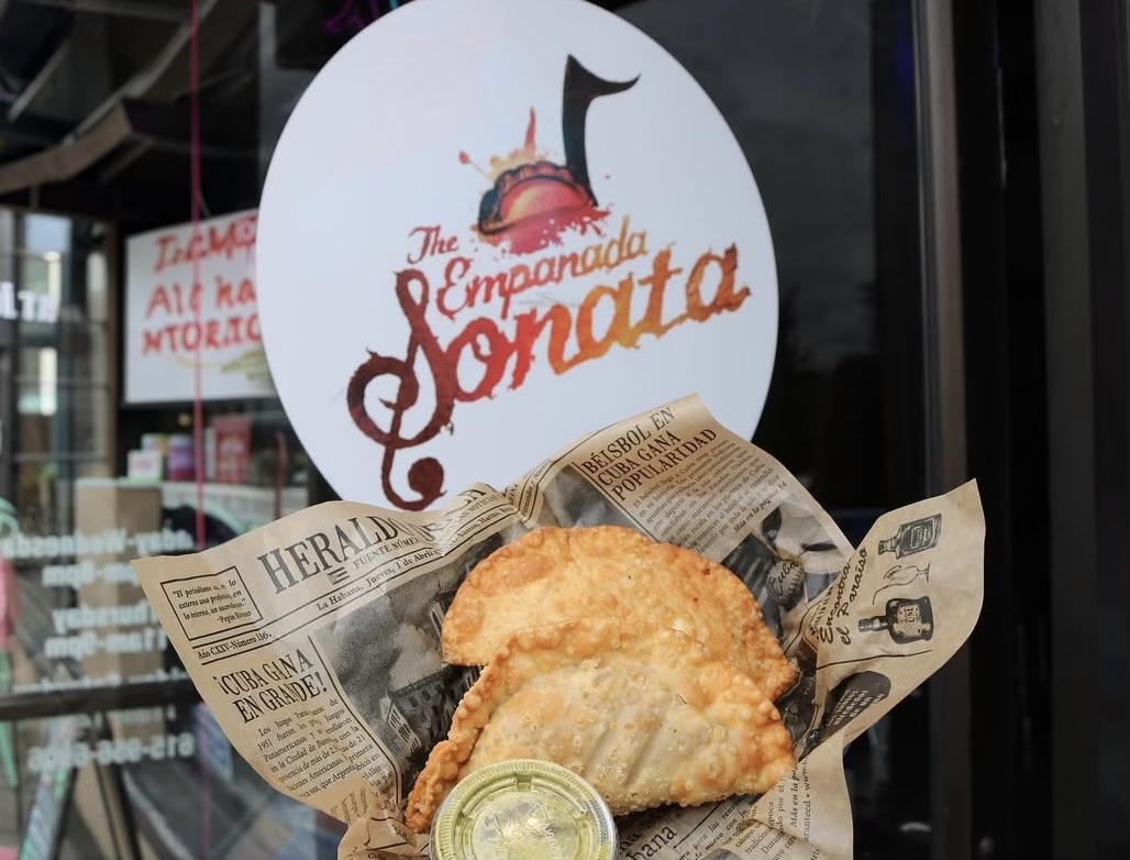 Two fried empanadas on newspaper-lined tray with a small cup of dipping sauce, in front of a sign for The Empanada Sonata.