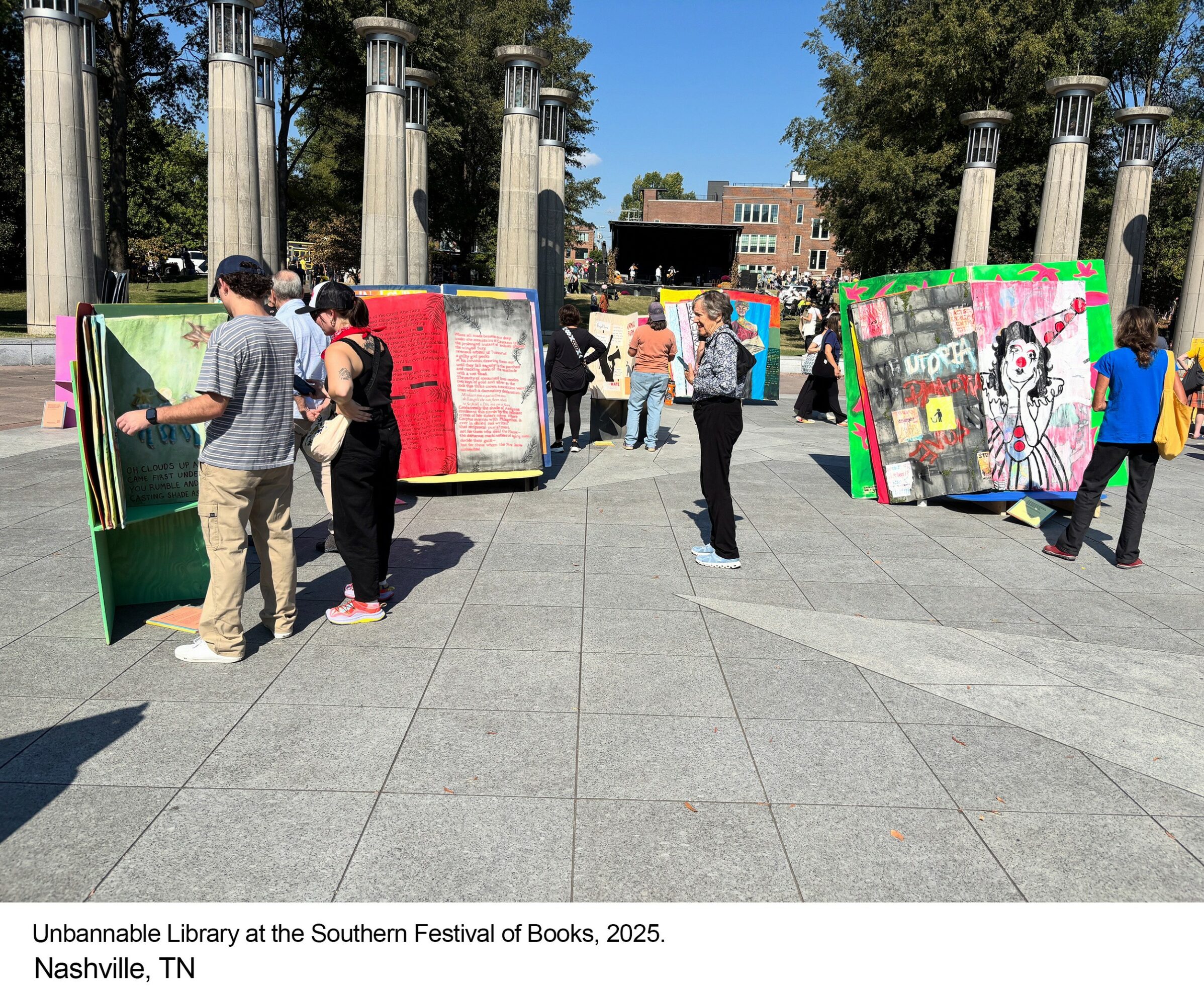 People browsing colorful book displays at an outdoor festival with columns and trees in the background.