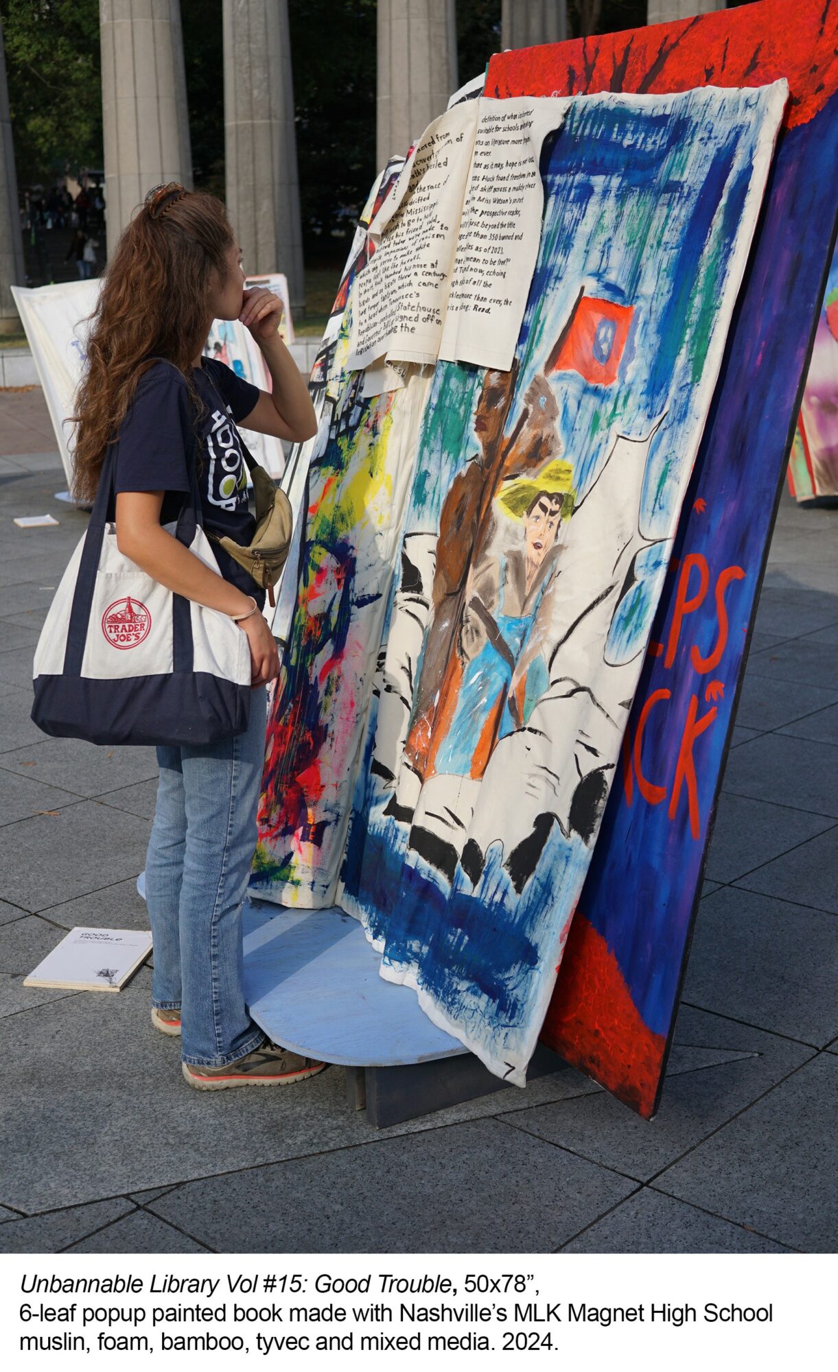 Young girl with curly hair, wearing a dark T-shirt and jeans, looks at colorful artwork displayed outdoors.