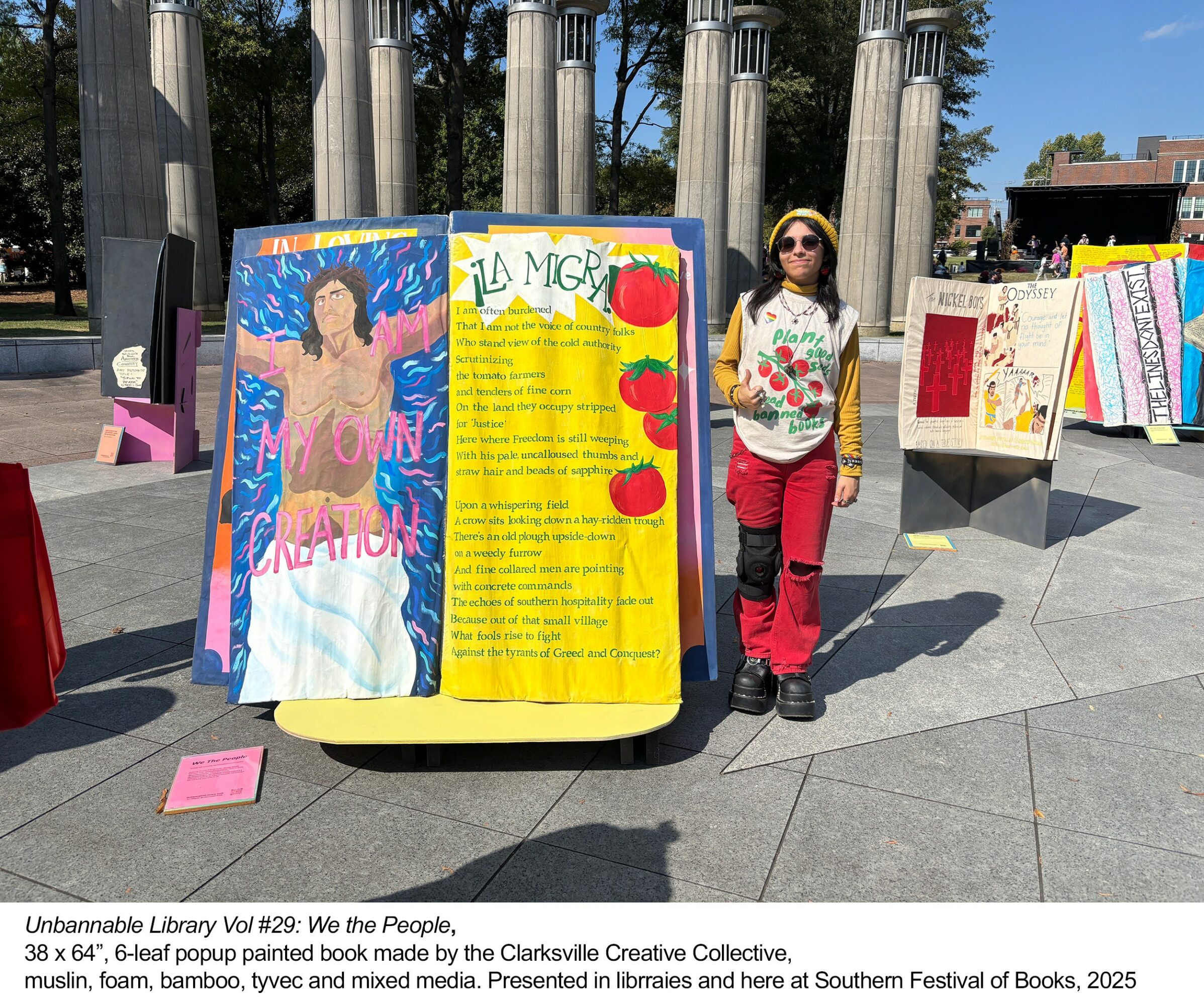 Person standing next to colorful display boards with artwork and text outdoors, columns and trees in background.