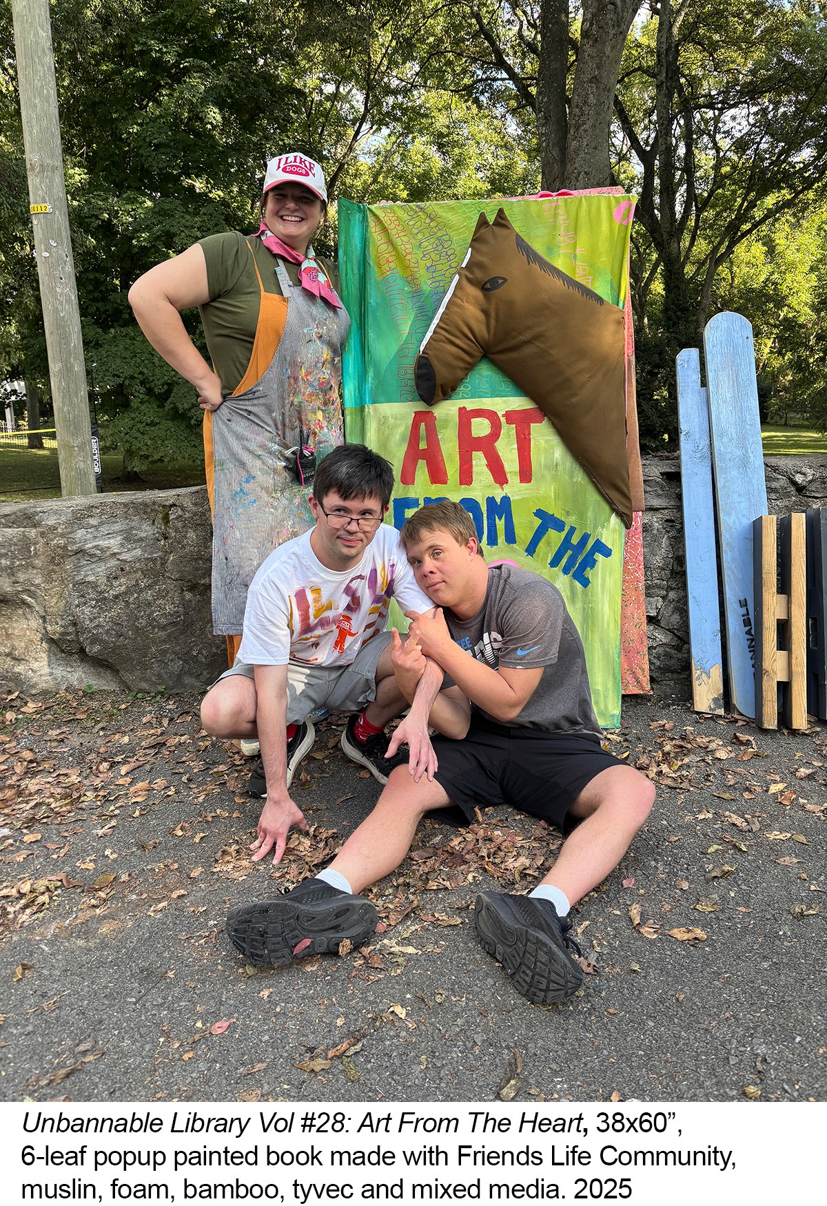 Two boys sitting on the ground in front of a colorful art display, with a woman standing behind them outdoors.