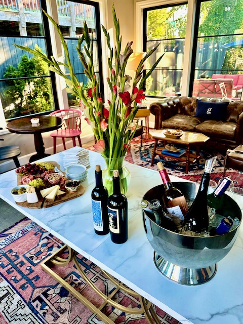 Vase with tall red and yellow flowers on a table, bottles, and a bucket with wine bottles in a living room.