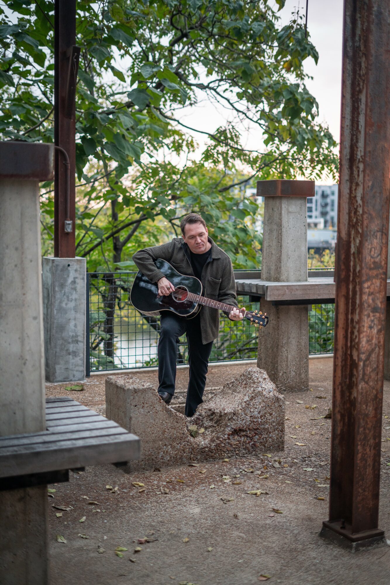 Man playing guitar outdoors near trees and wooden structures, with a cityscape in the background.