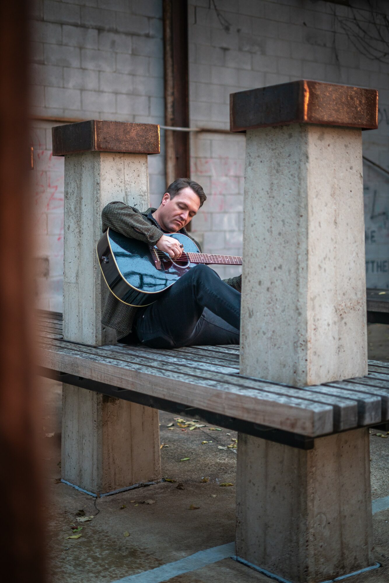 Man sitting on wooden platform playing guitar under concrete structure in outdoor setting.