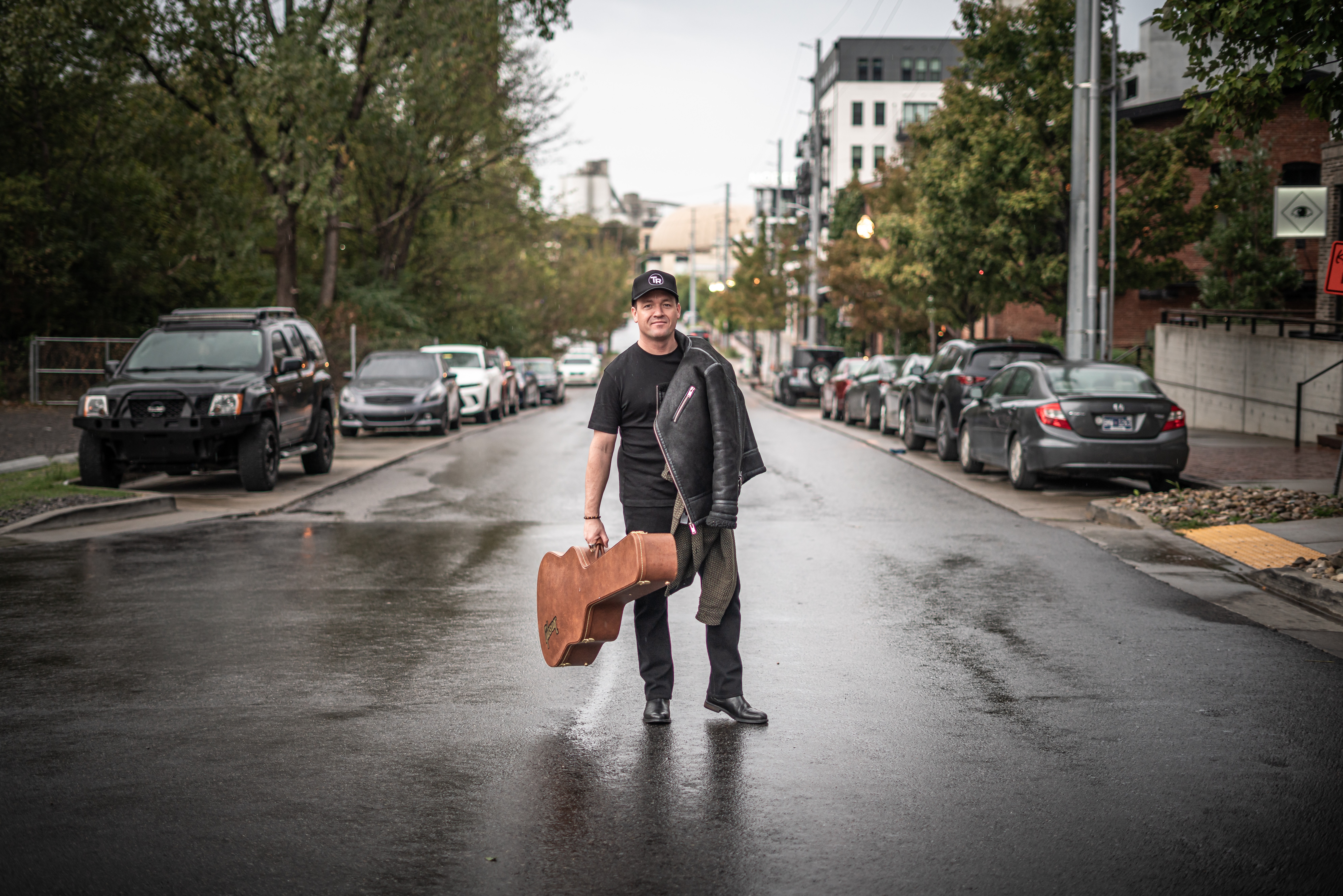 Person walking on wet street holding a guitar case, surrounded by parked cars and trees, urban setting.