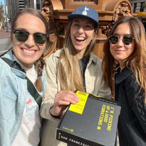 Three women smiling outdoors, one holding a book, wearing sunglasses and casual clothing, with a wooden structure in the background.