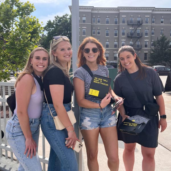 Four women standing outdoors on a sunny day, smiling, holding books, with trees and a building in the background.