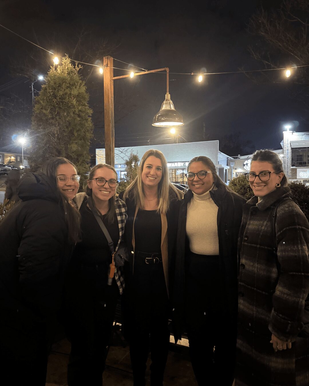 Group of five women standing outdoors at night, smiling, with string lights overhead and a building in the background.