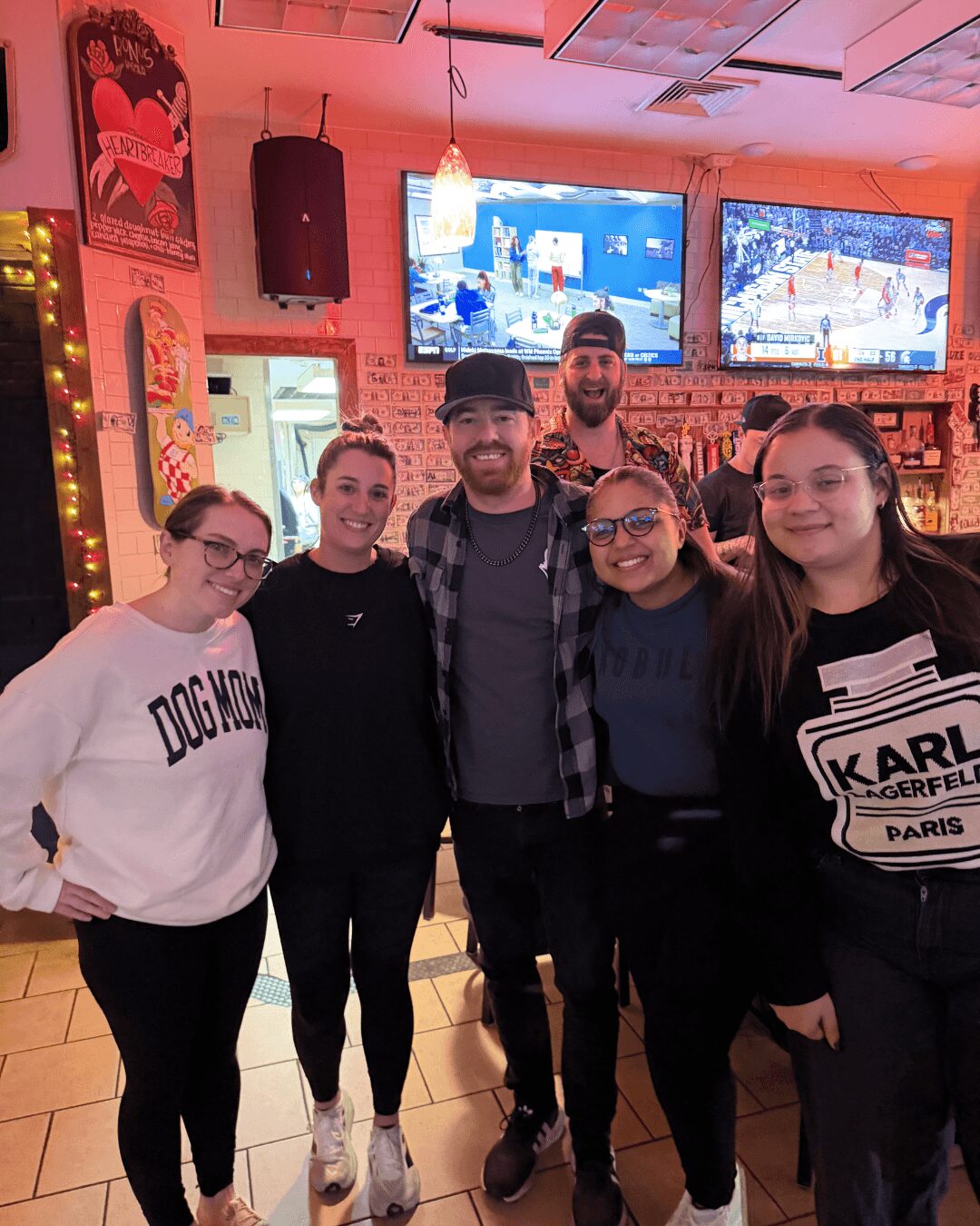 Six people standing together in a bar, smiling, with TVs and colorful decor in the background.