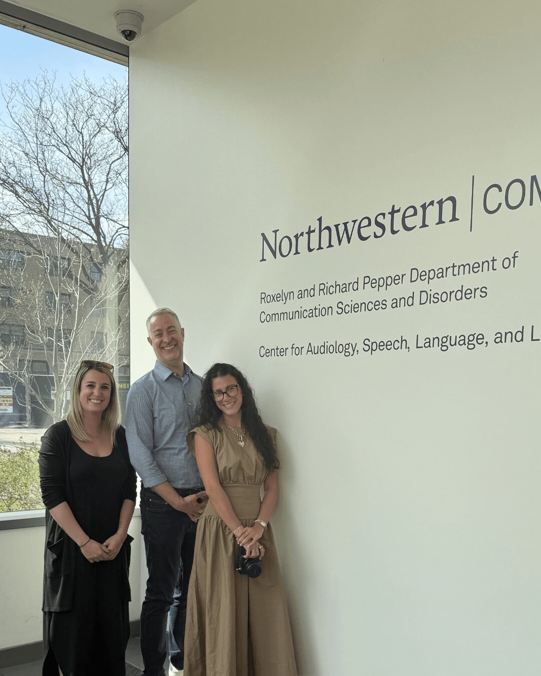 Three people standing indoors near a wall with text, smiling, with a window showing trees outside.