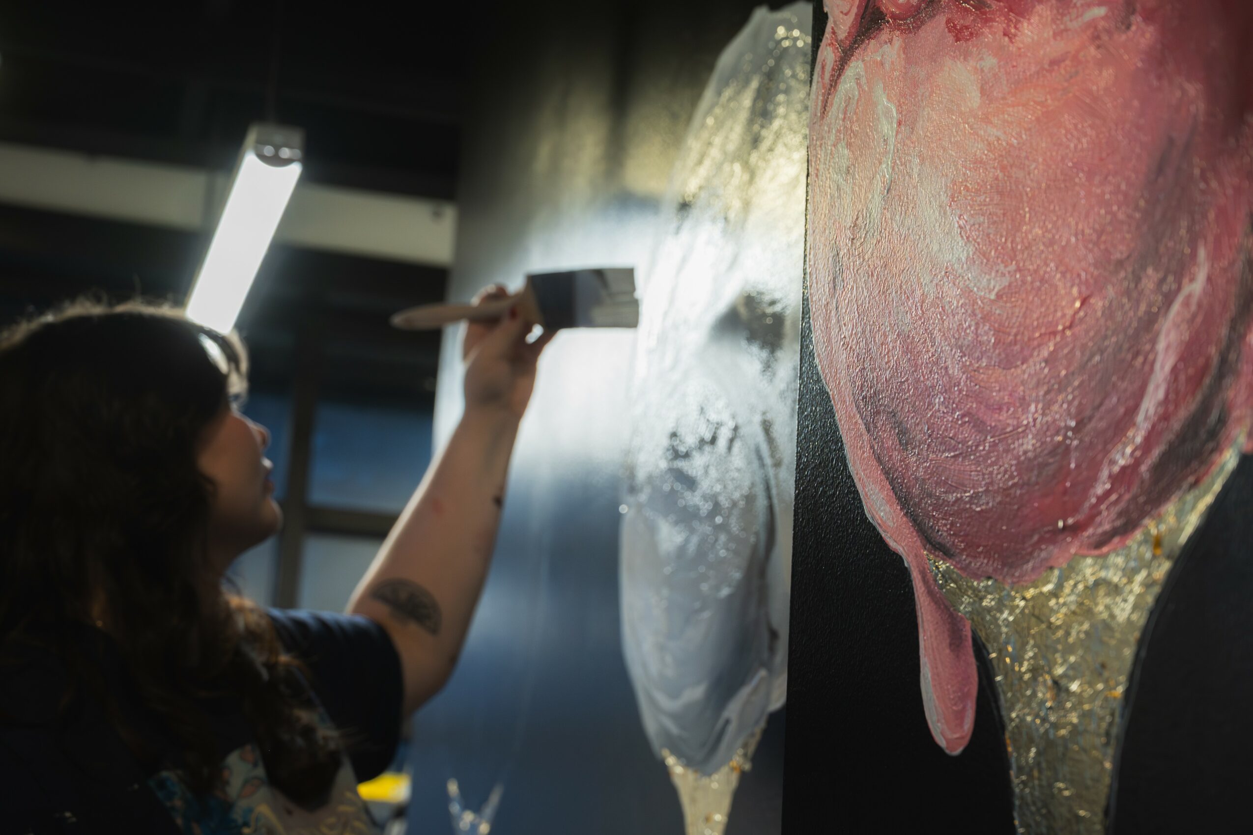 Person painting a large, colorful artwork on a wall in an indoor space, with a light fixture overhead.
