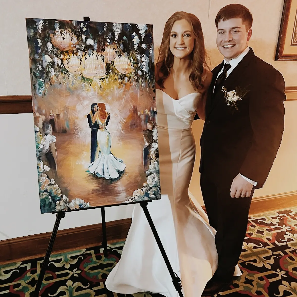A bride and groom stand next to a wedding painting on an easel, smiling indoors with a patterned carpet.