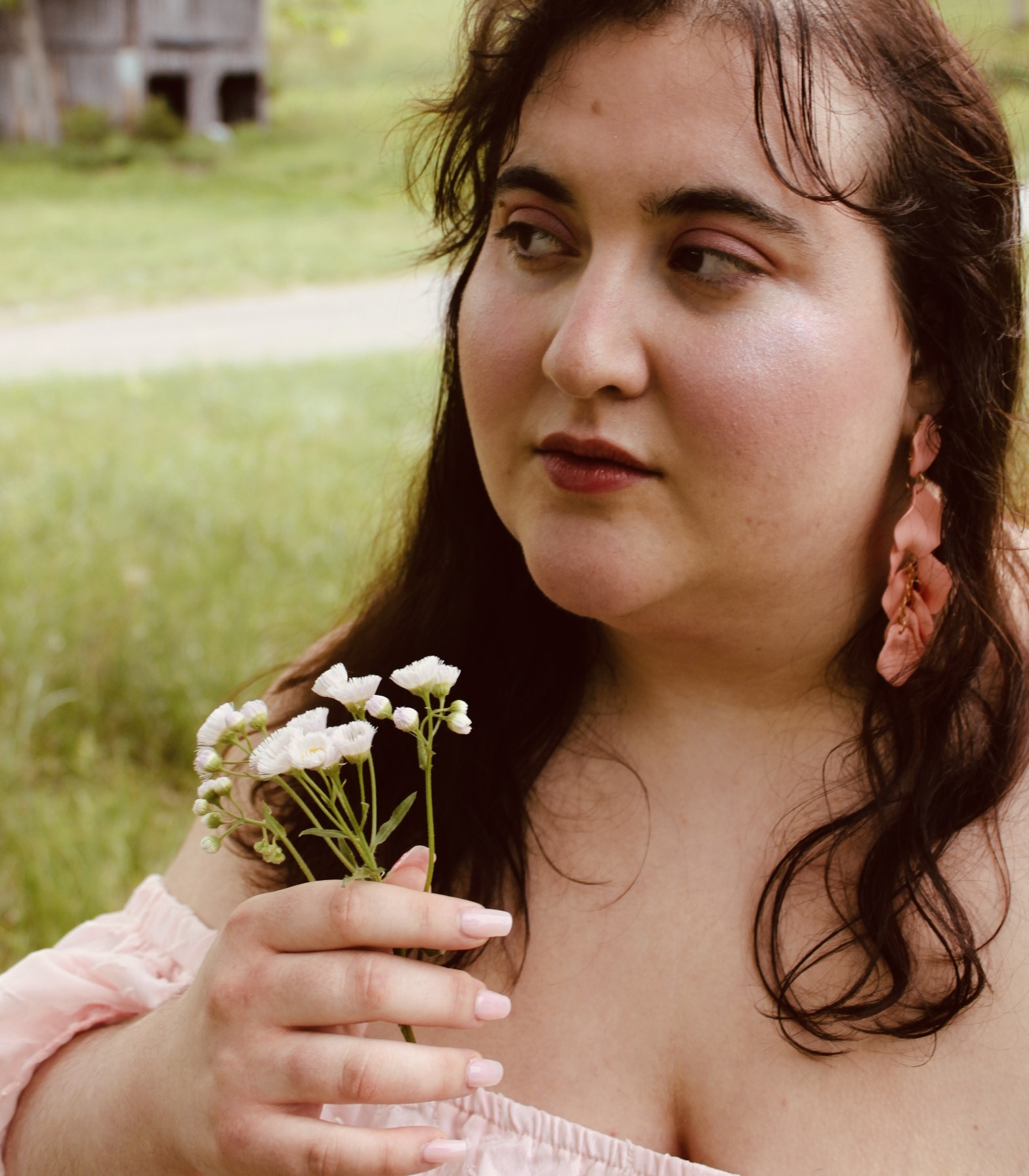 Young woman with dark hair holding small white flowers outdoors, wearing pink earrings and a pink top.