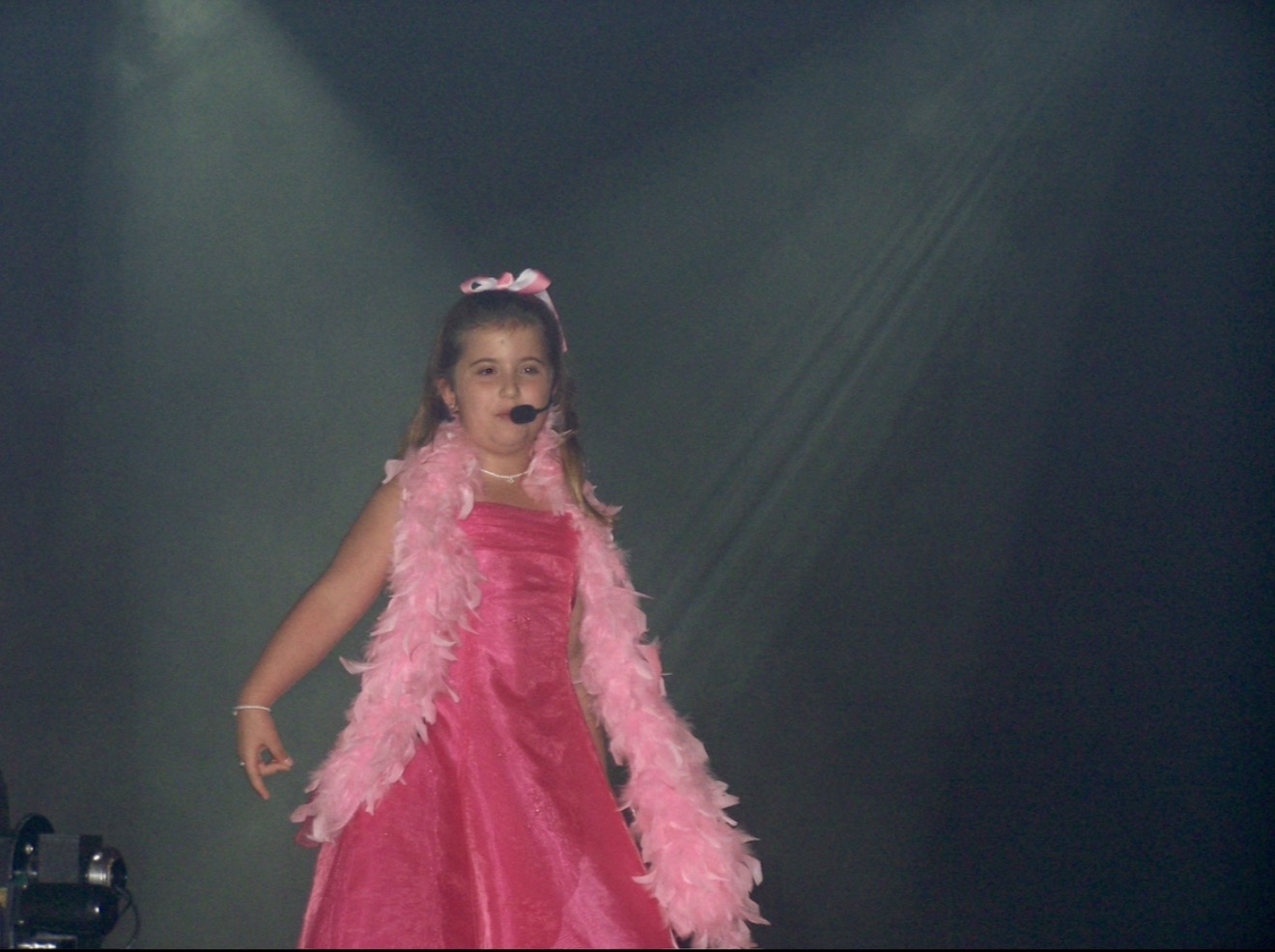 Young girl in pink dress and feathered boa on stage with dark background.