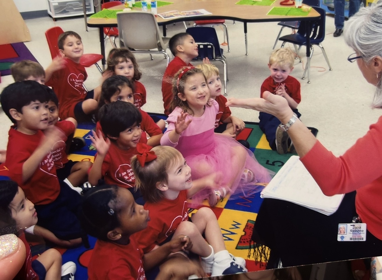 Group of children sitting around a teacher reading a book, in a colorful classroom setting.